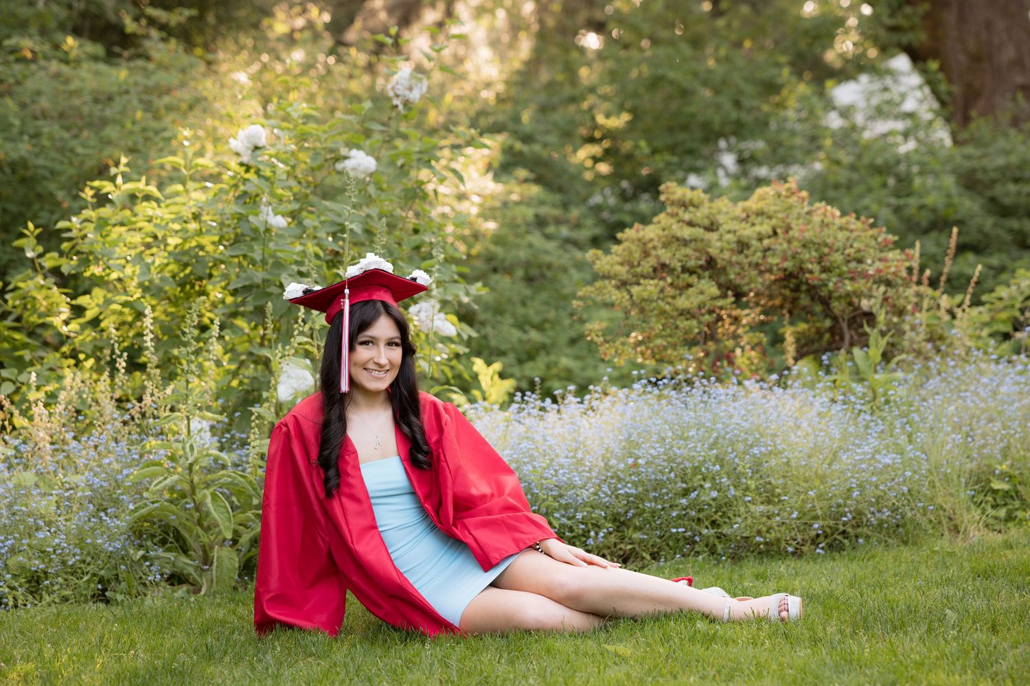 Young woman in red graduation cap and gown sitting on grass surrounded by lush green trees and wildflowers.