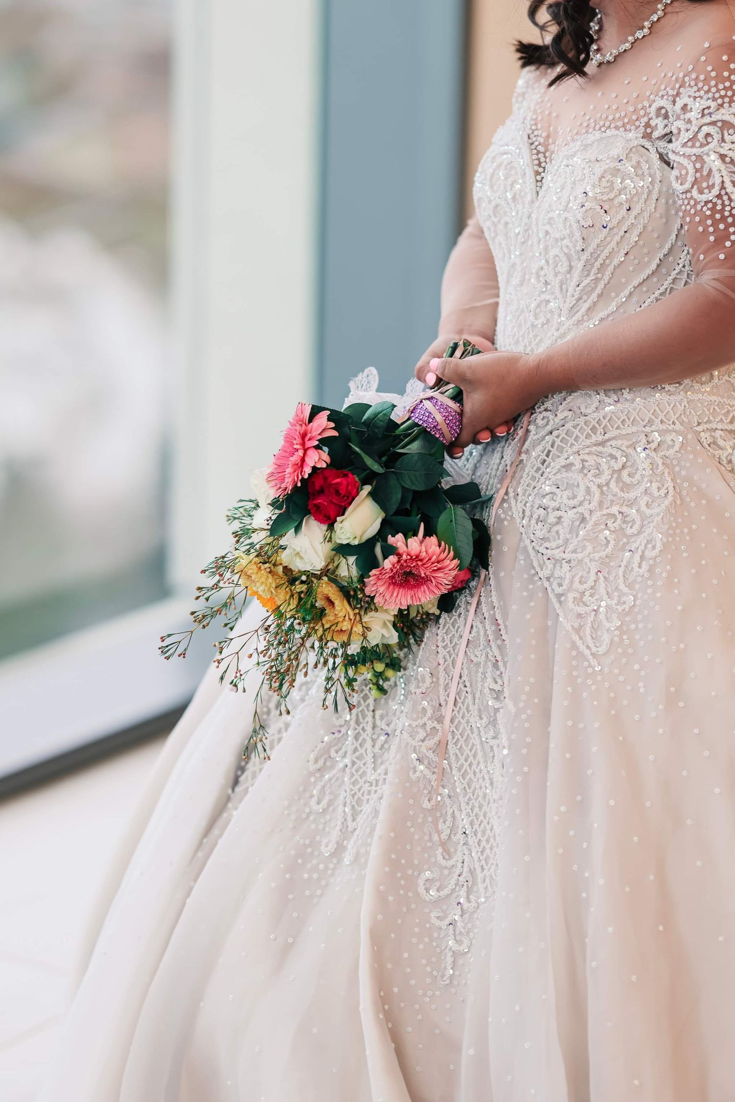 Bride in beaded lace wedding gown holding colorful bridal bouquet with pink gerberas, roses, and greenery.
