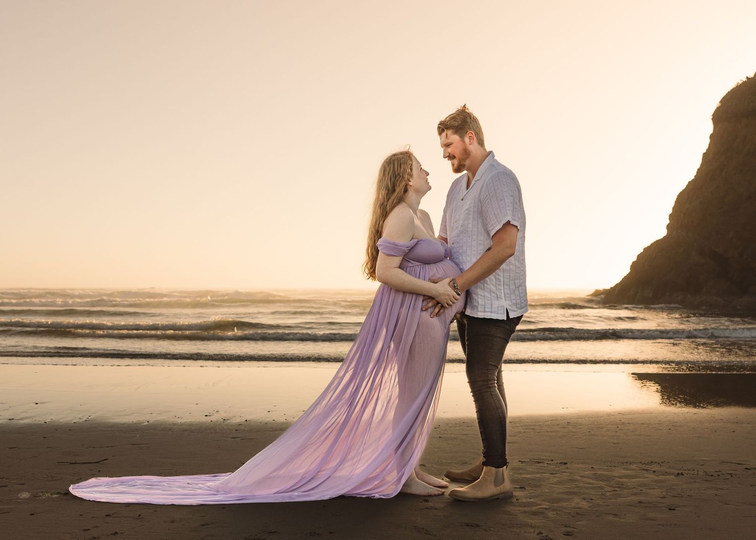A couple embraces on a sandy beach at sunset with cliffs and ocean waves in the background.