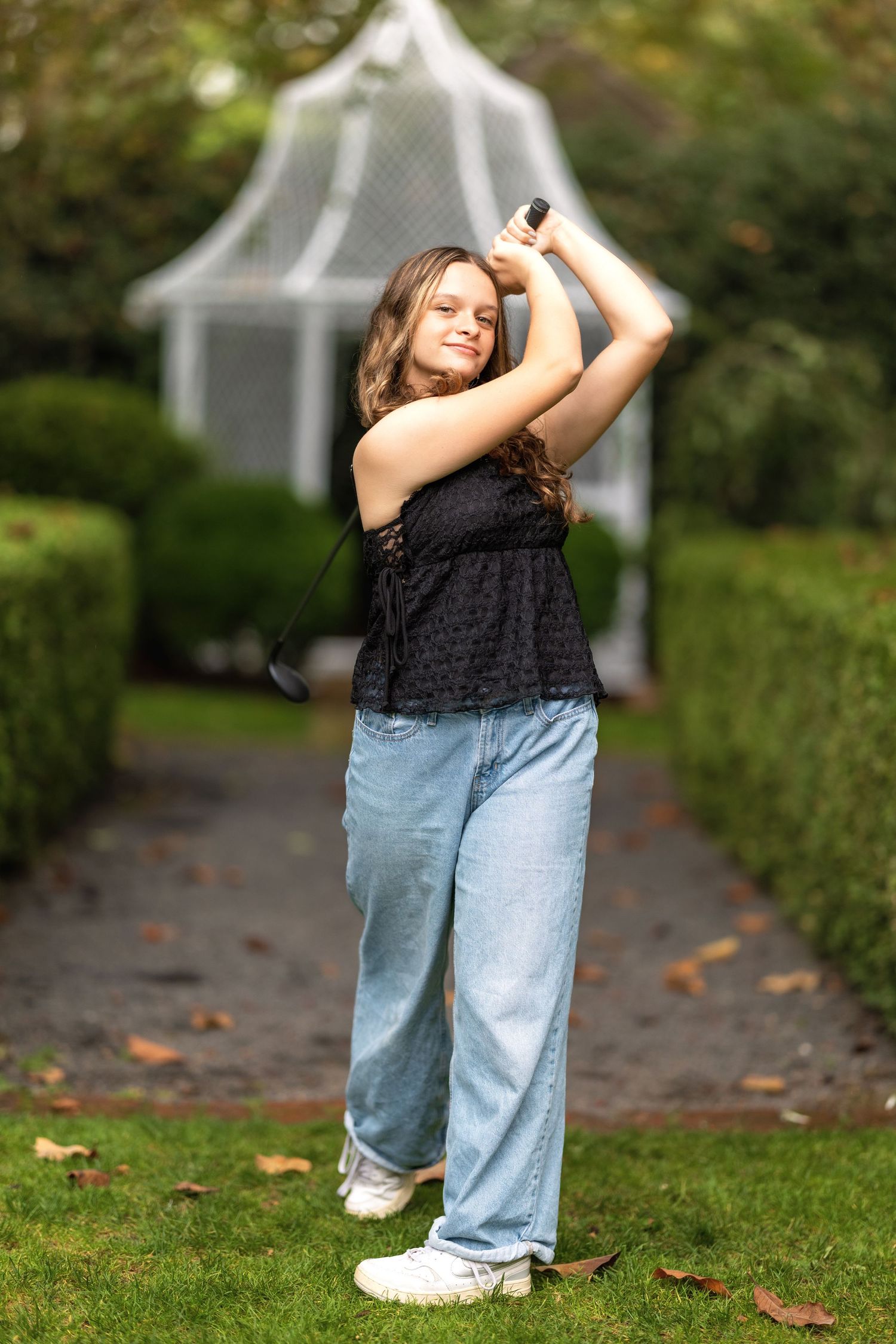 A young girl in jeans and black top swings a golf club in a lush green garden with a white gazebo.