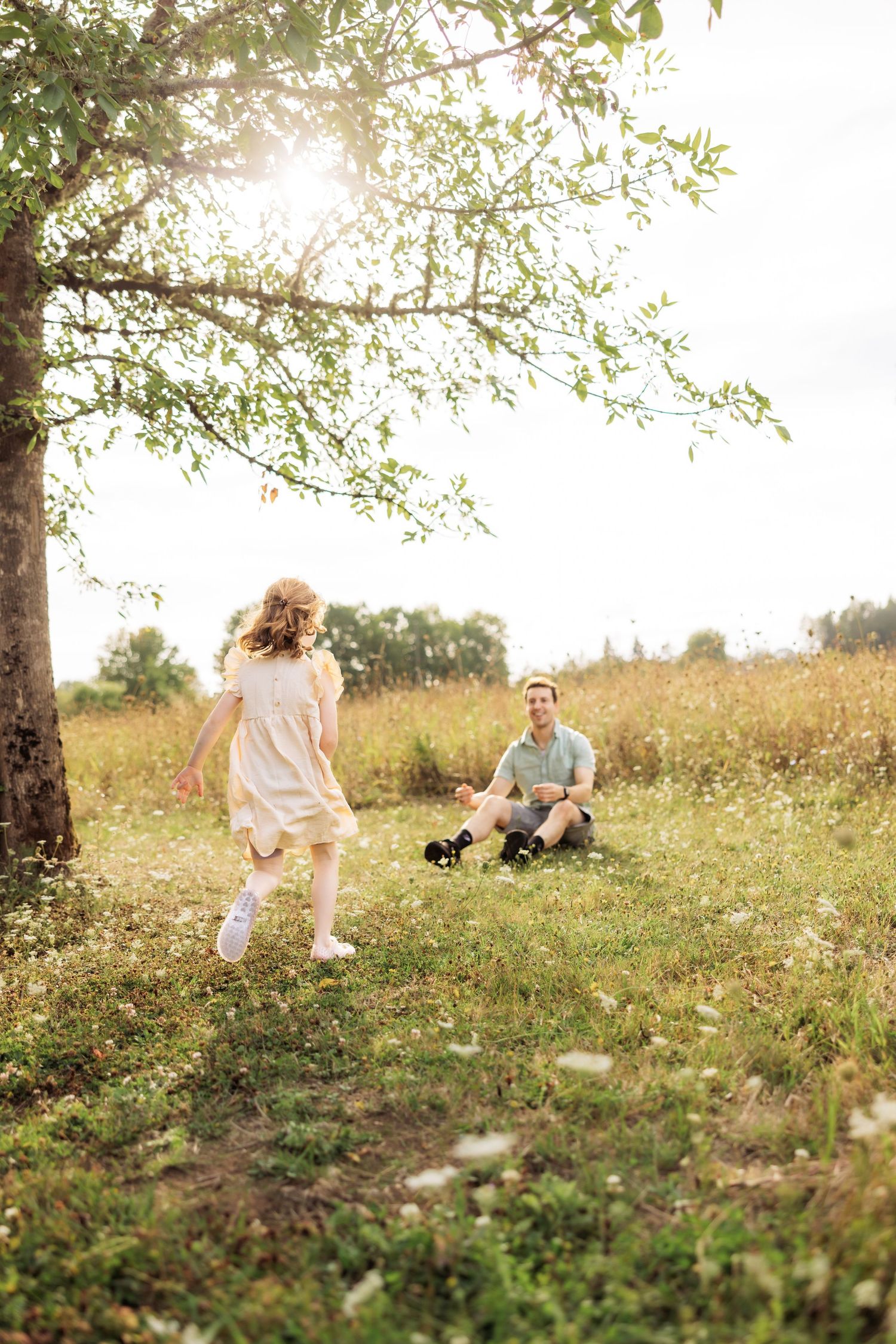 A young girl in a white dress runs toward her father sitting on a sunny green meadow beneath a leafy tree.
