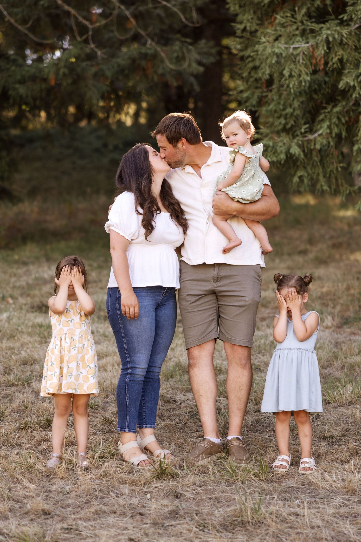 Family of five outdoors, parents kissing while holding toddler, two young girls covering their faces with hands.