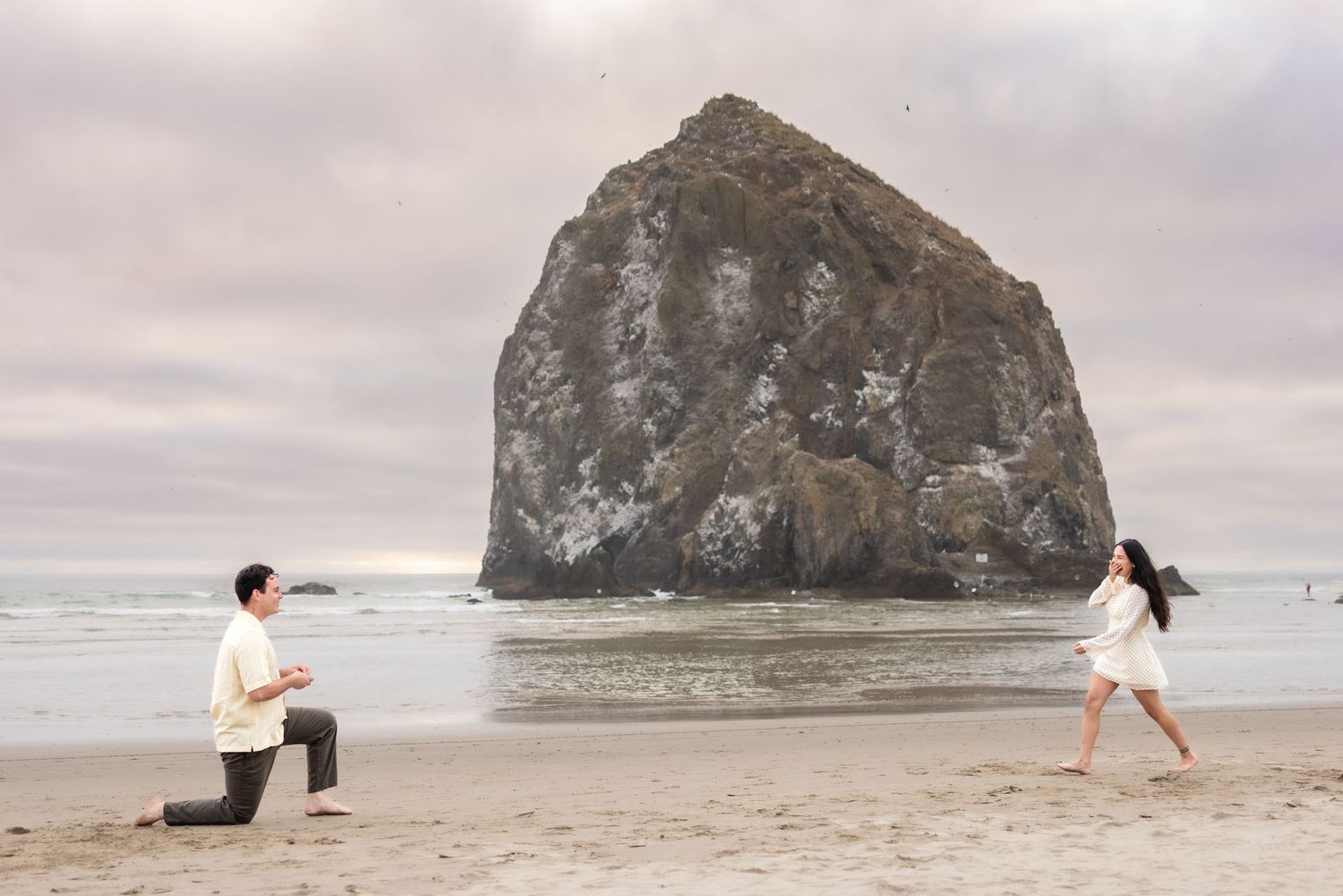 Man proposes on one knee to woman on sandy beach with iconic Haystack Rock at Cannon Beach Oregon in background.