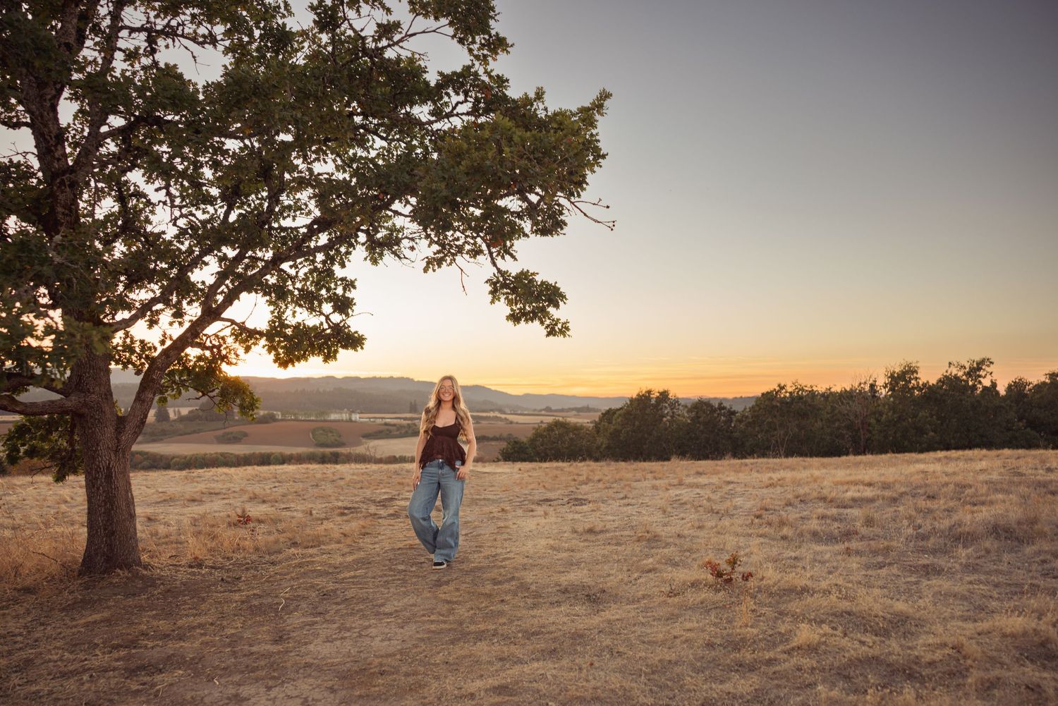 Woman standing in golden field at sunset near oak tree with rolling hills in background.