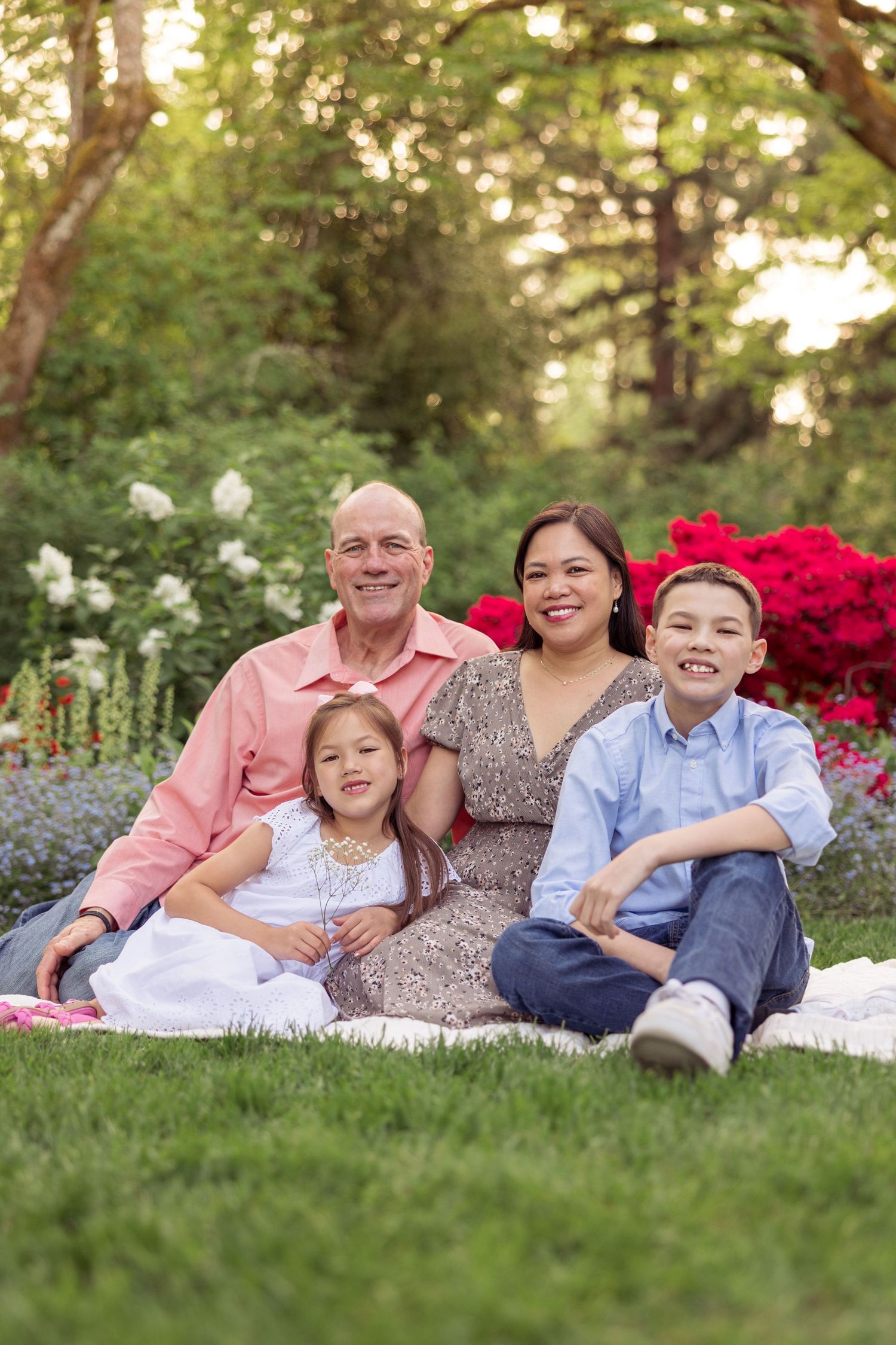 Family relaxing together on a blanket in a garden with blooming red azalea bushes in the background during sunset.