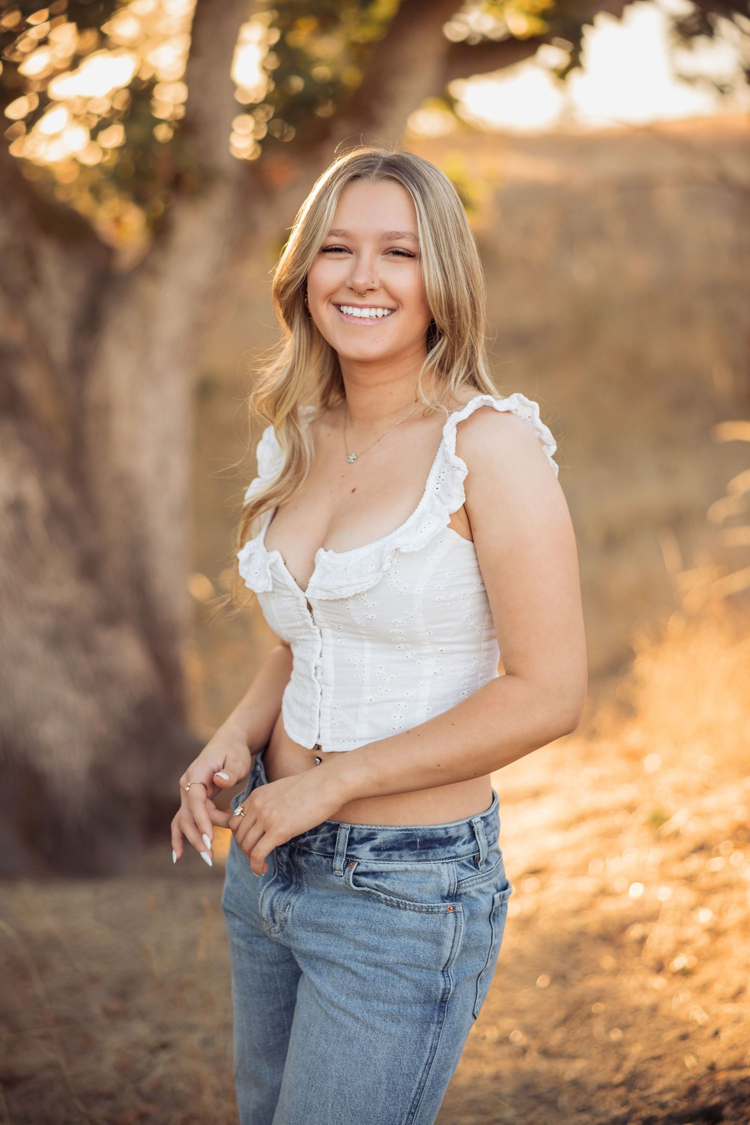 A person in a white ruffled top and blue jeans poses outdoors during golden hour sunset lighting.