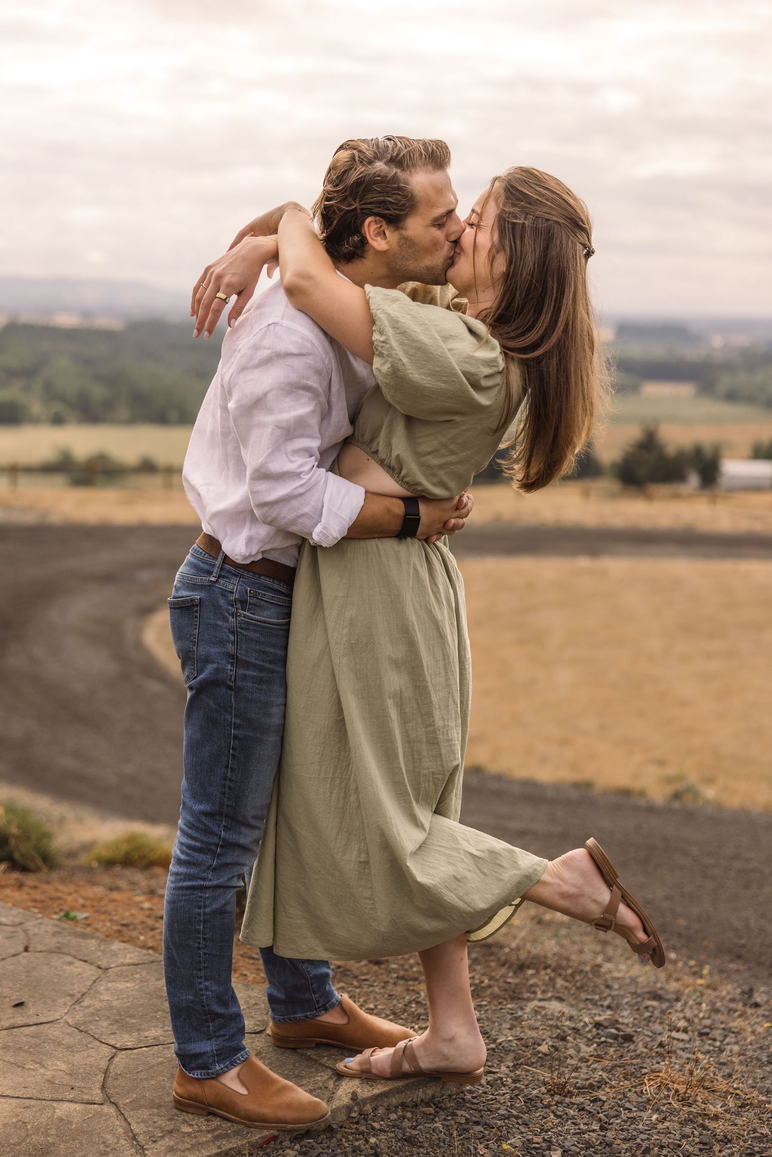 A couple shares a romantic moment on a hilltop overlooking a scenic rural landscape at sunset.