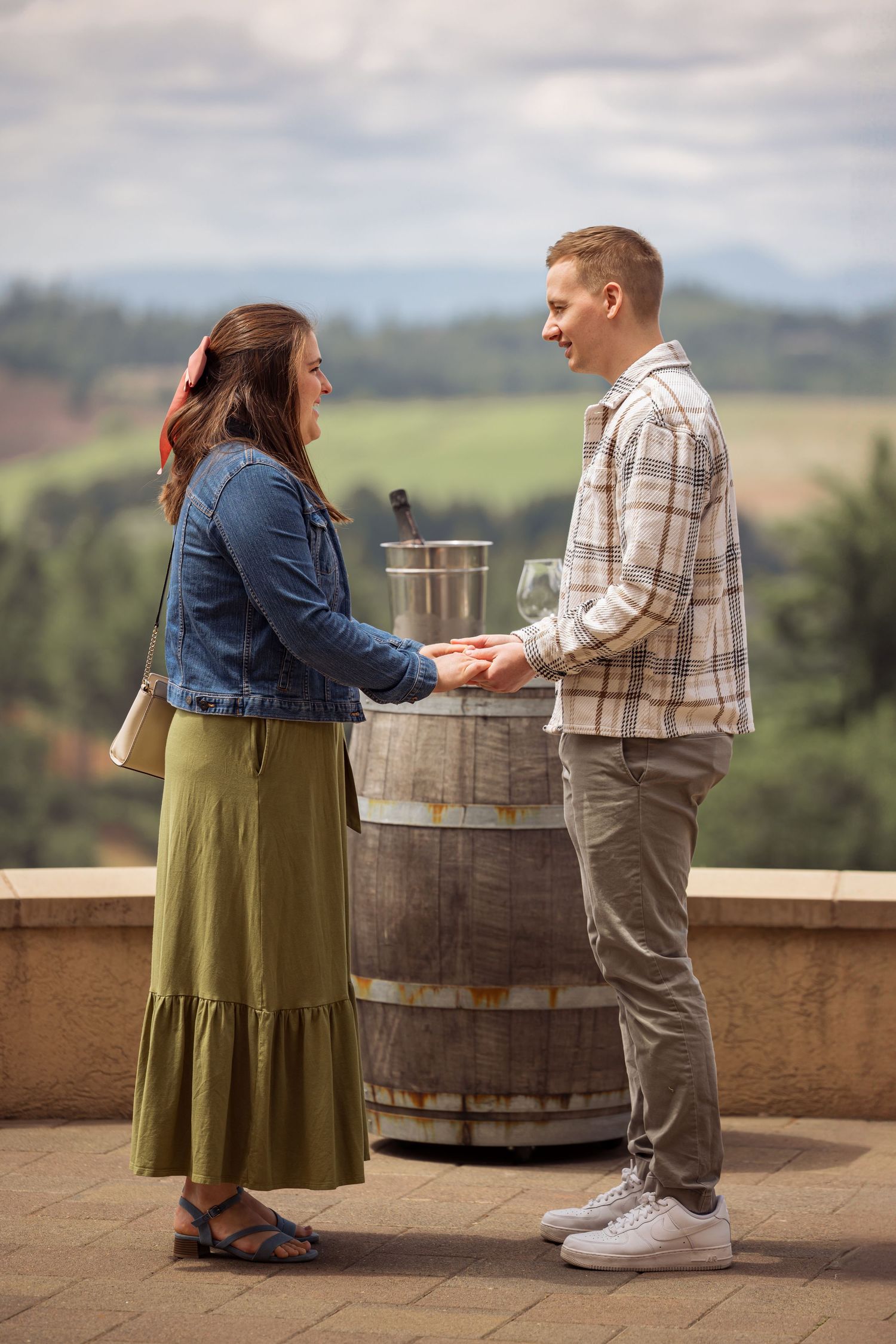 Two people share an intimate moment on a winery balcony overlooking scenic hills with wine barrels in the background.