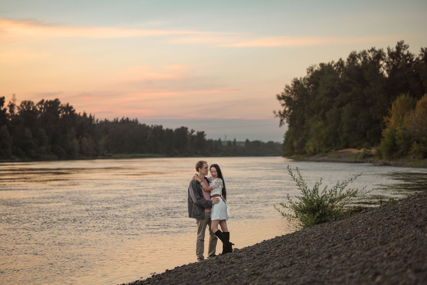 Couple embracing by a river at sunset with lush trees and golden sky in the background.
