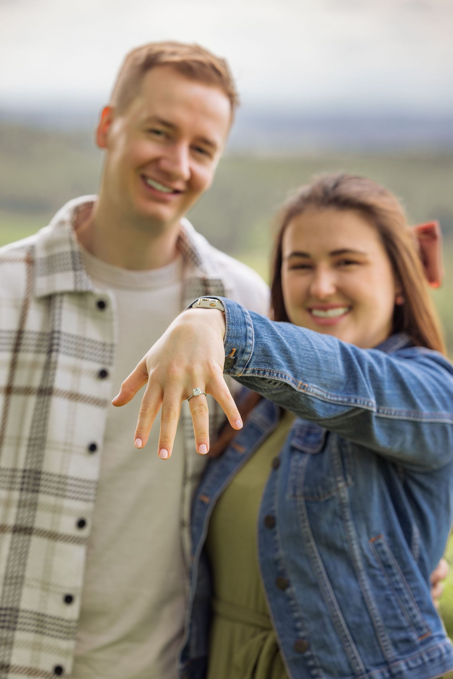 A newly engaged couple showing off an engagement ring while celebrating outdoors with wine.