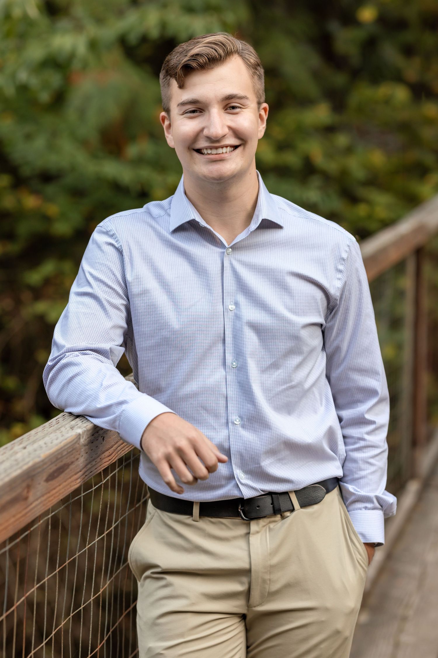 Professional portrait of someone in a light blue dress shirt and khaki pants standing casually on a wooden bridge outdoors.