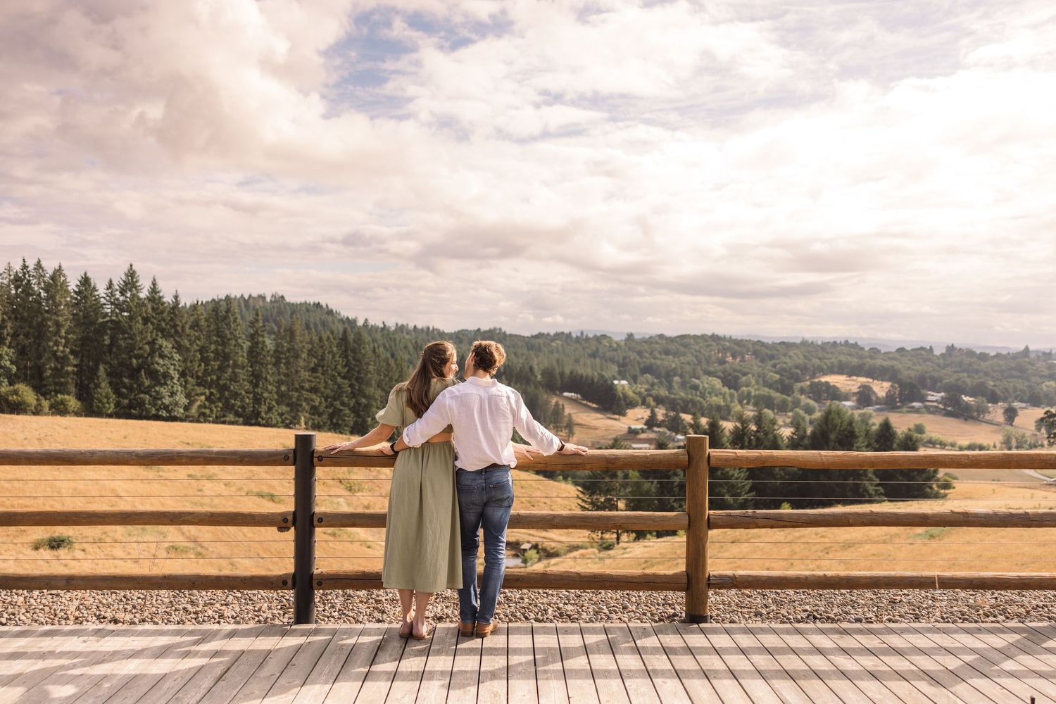 A couple stands together on a wooden deck overlooking rolling hills and farmland on a cloudy day.