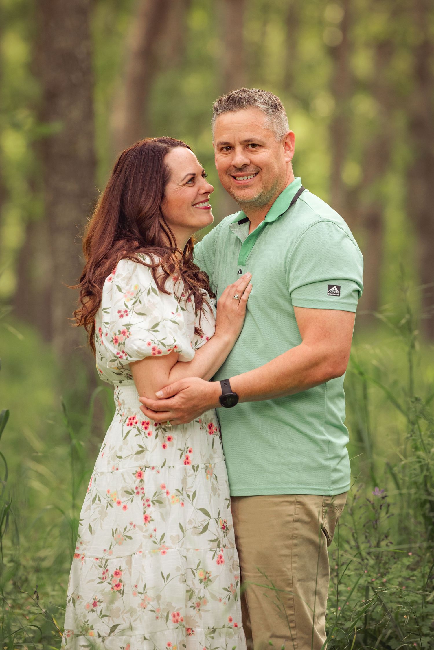 A couple shares a loving moment outdoors standing together in a wooded area, she in a floral dress and he in mint green polo.