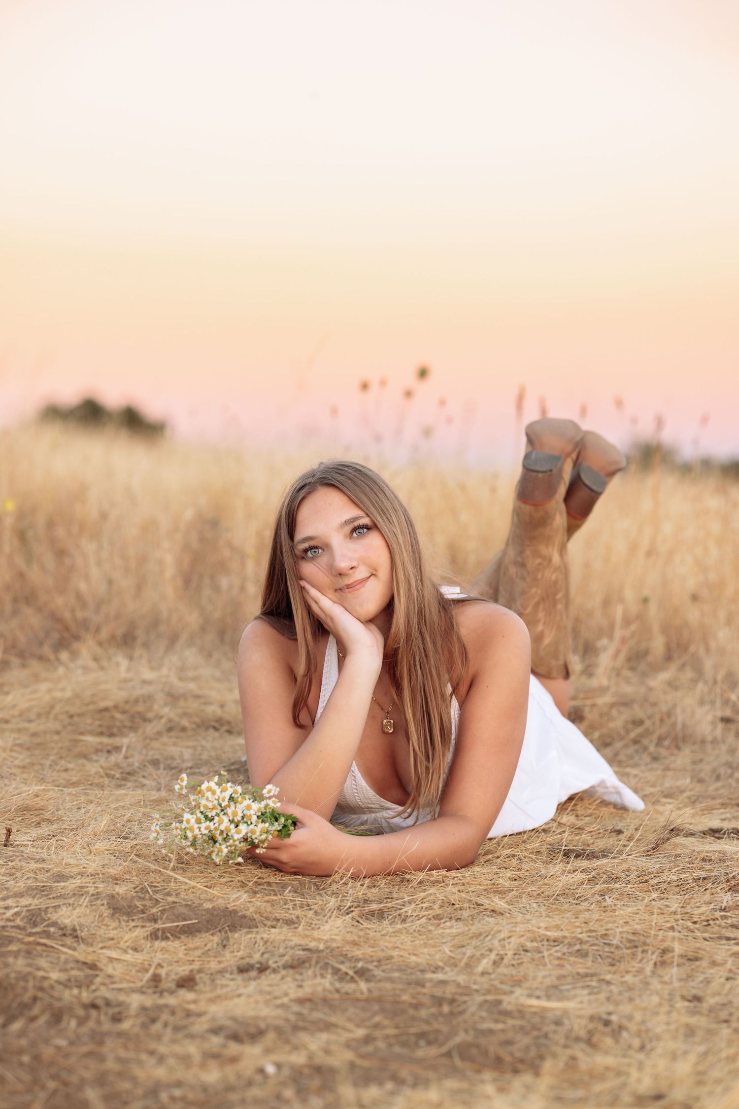 Young woman in white dress lying in golden field holding wildflowers at sunset.