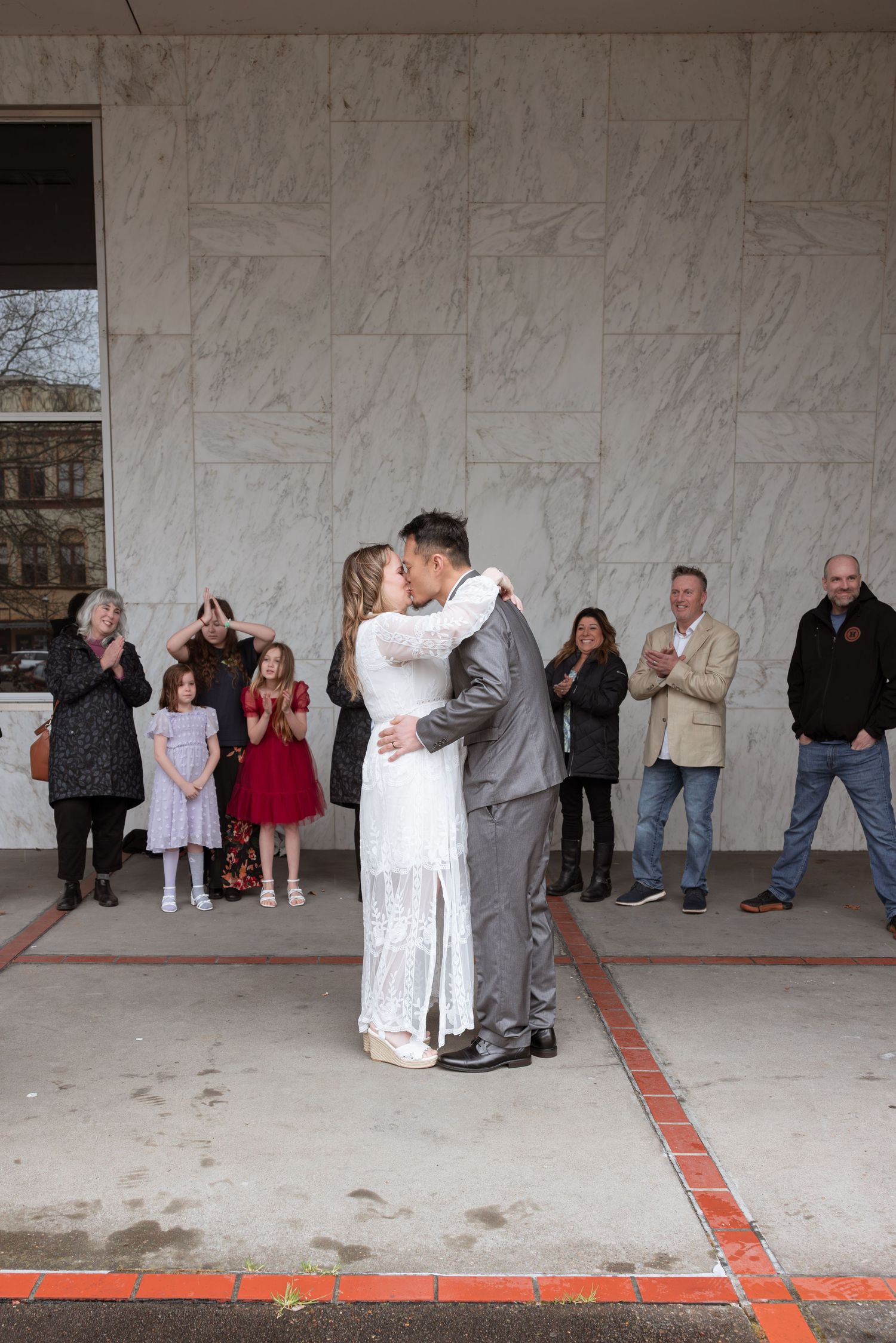 Couple shares first kiss at outdoor courthouse wedding ceremony while guests applaud and children watch in festive attire.