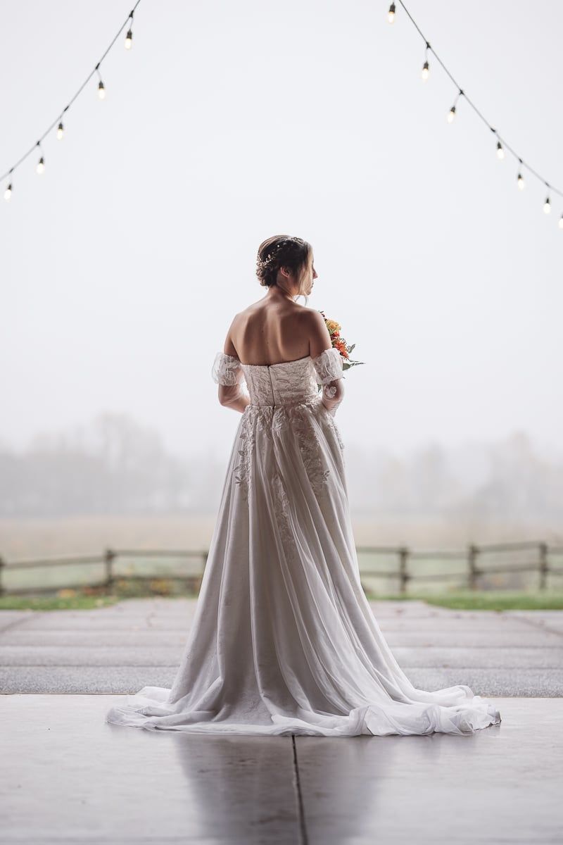 Bride in elegant off-shoulder white lace wedding gown with train stands on patio gazing at misty countryside landscape.