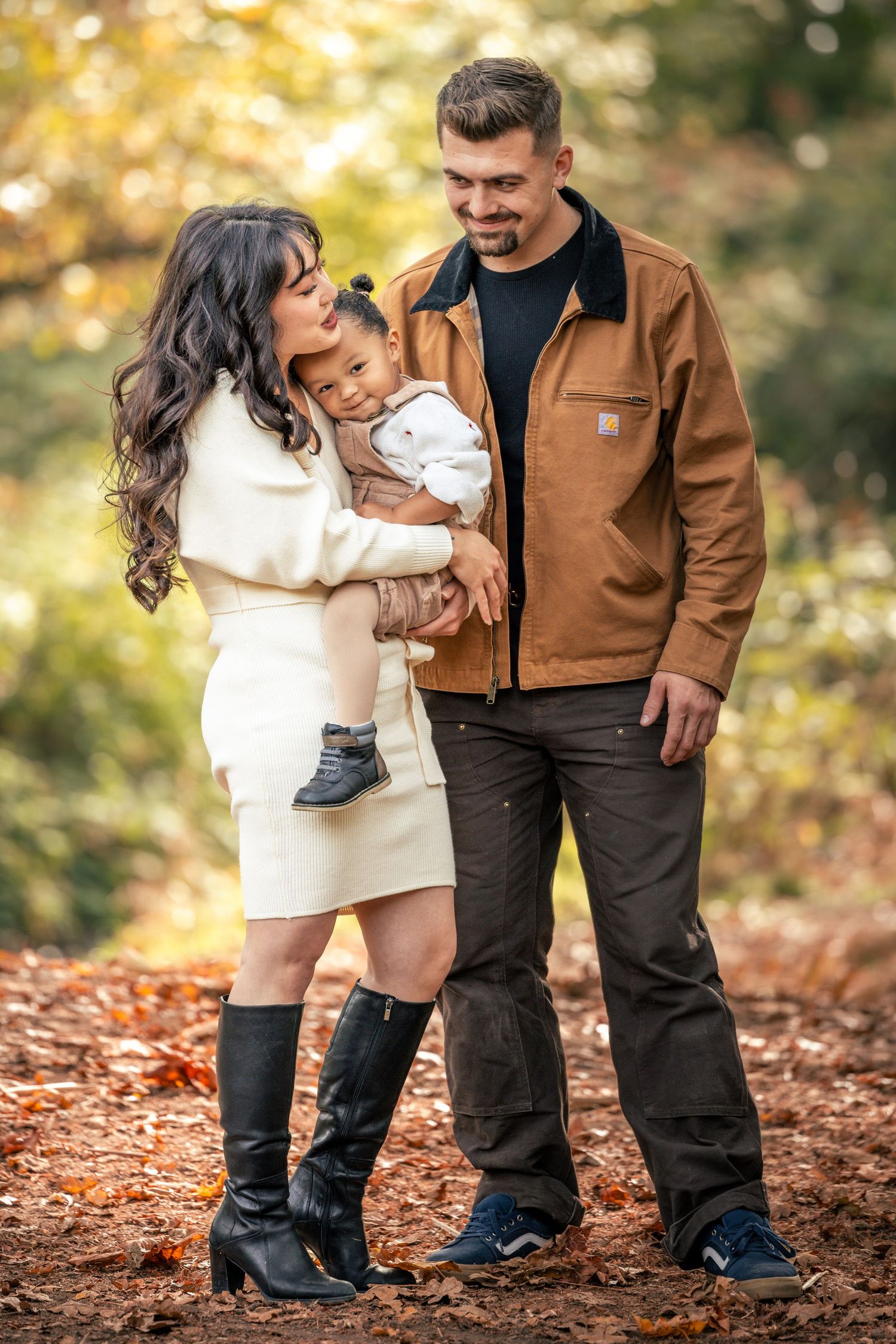 Family portrait in autumn setting wearing contrasting white and brown outfits standing on fallen leaves in forest.