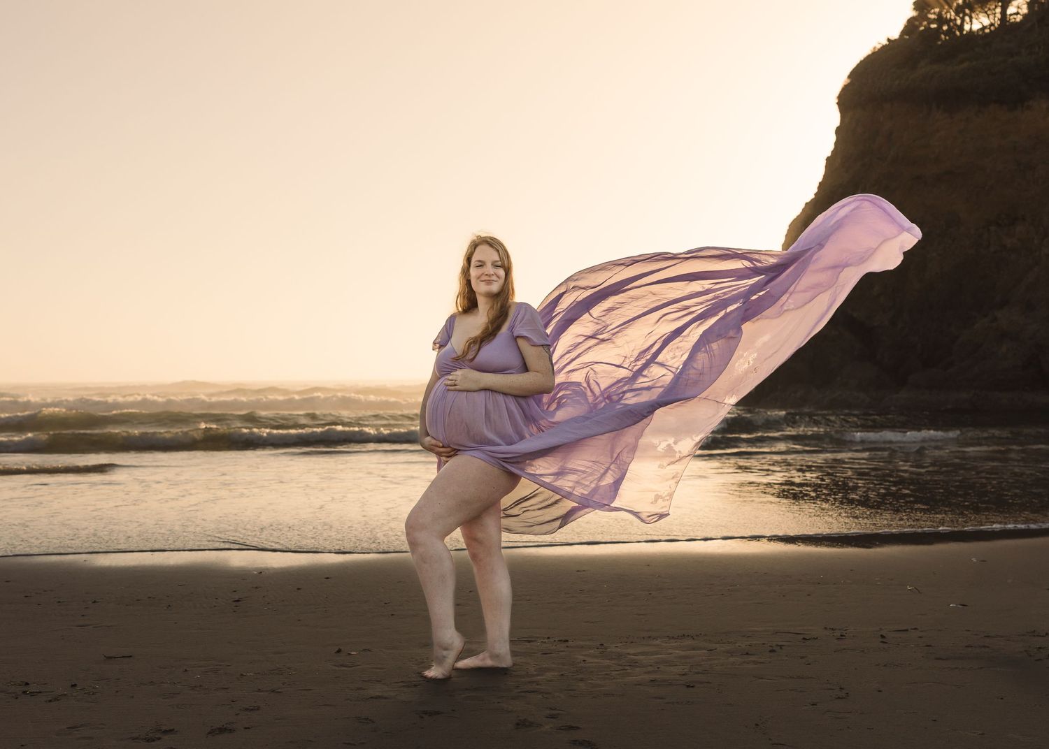 A flowing purple dress billows in the wind as someone poses on a beach at sunset against dramatic cliffs.