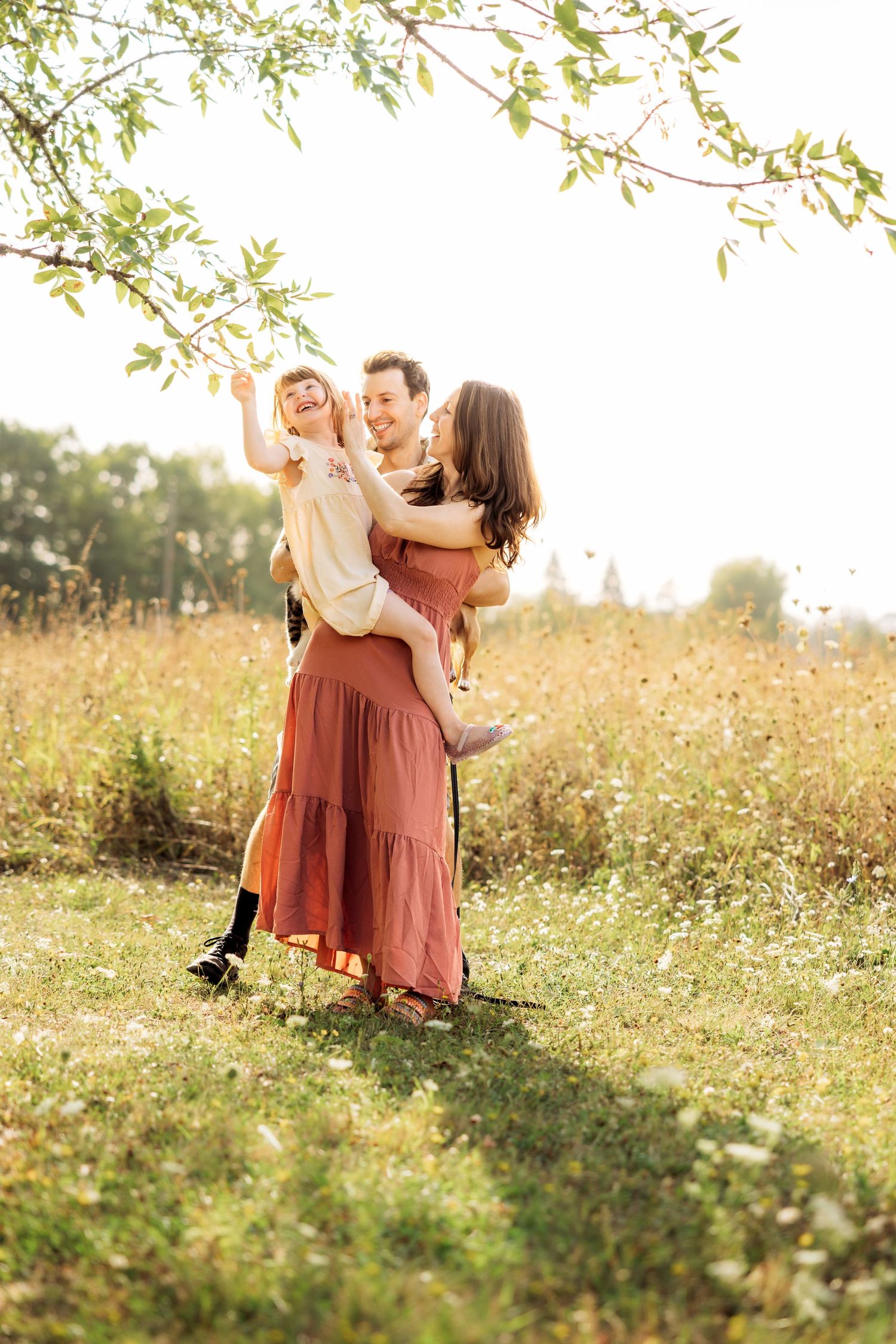 A joyful family of three plays outdoors in a sunny field, with dad lifting child while mom embraces them in warm light.