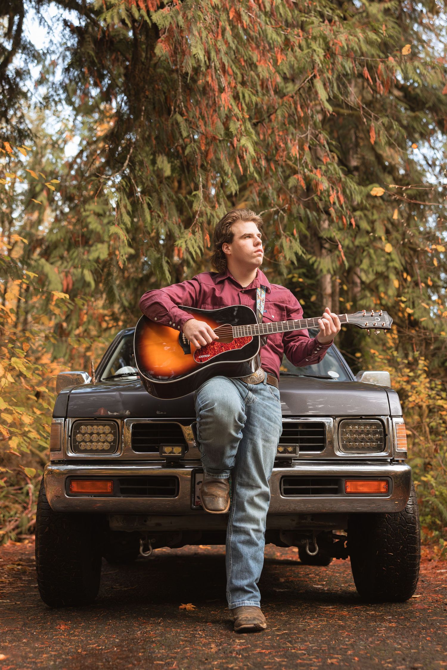 A musician sits on the hood of a vintage car playing acoustic guitar in an autumn forest setting.