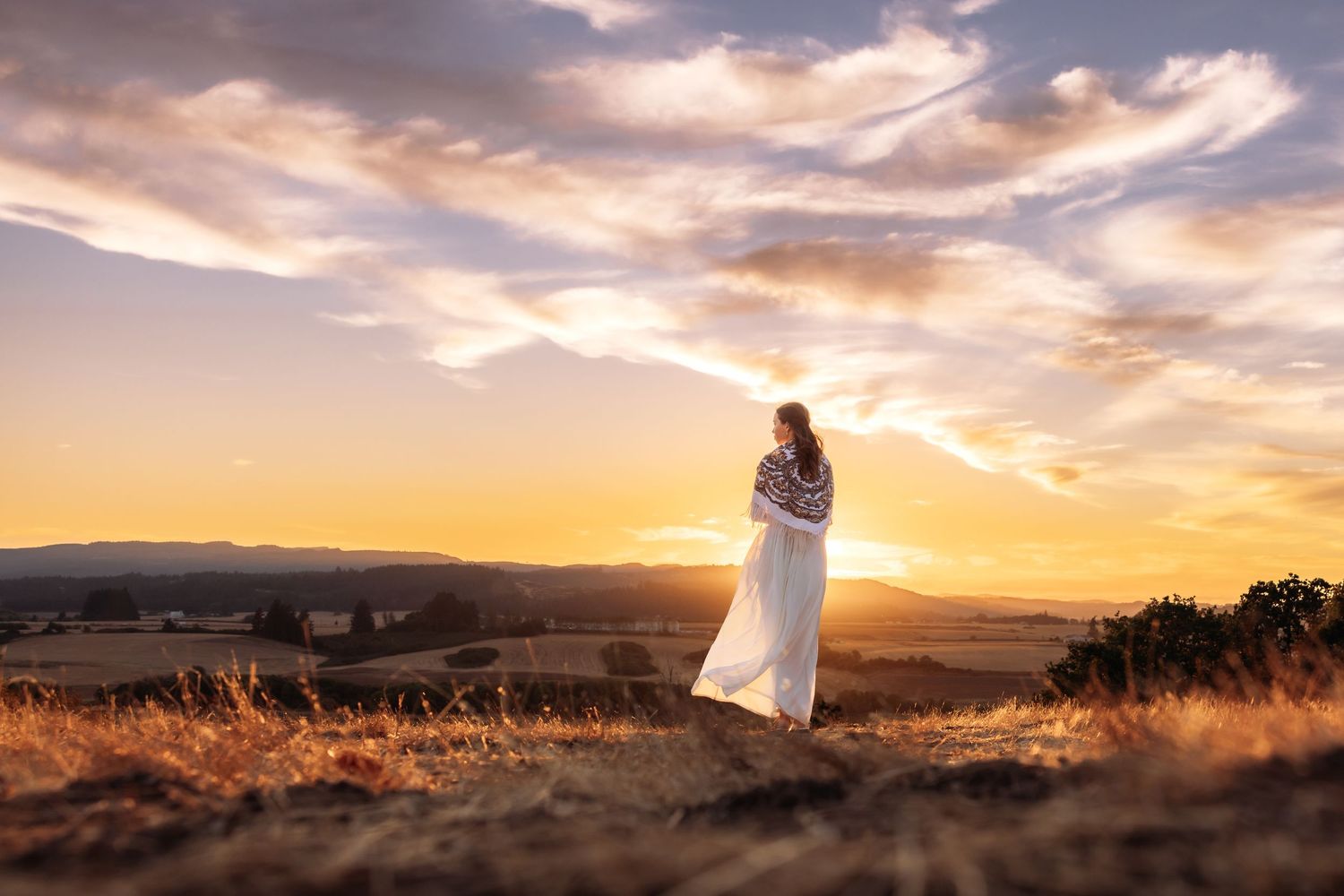 Woman in white dress standing on a rock at golden sunset with rolling hills and dramatic sky in background.