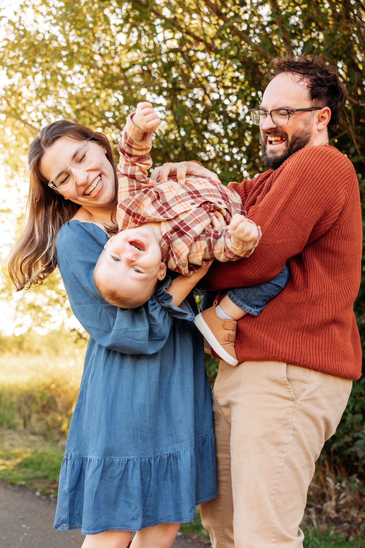 Happy family with parents laughing while playfully holding their baby outdoors in golden autumn light.
