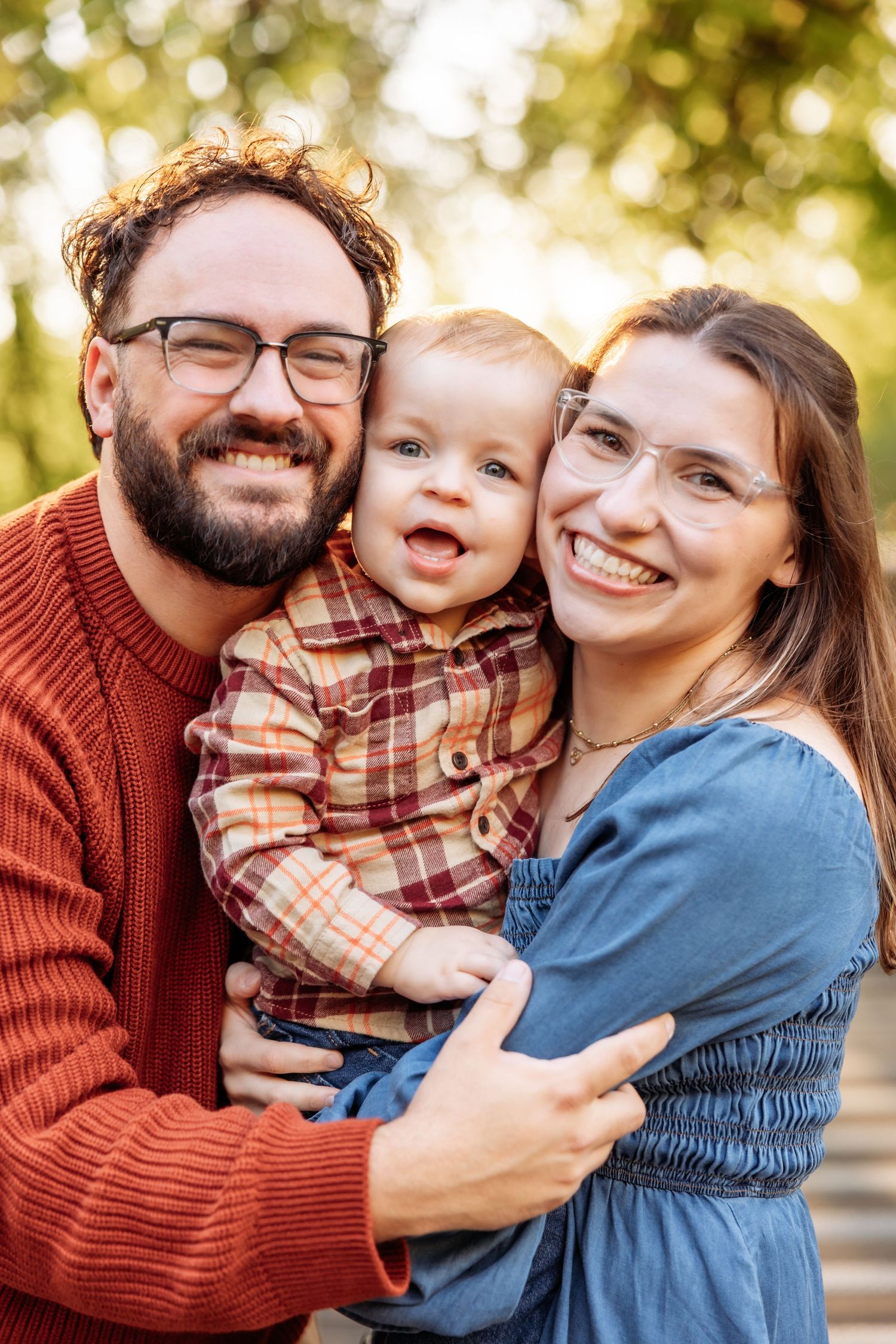 Happy family portrait with bearded dad, smiling mom in glasses, and adorable baby boy outdoors in autumn park.