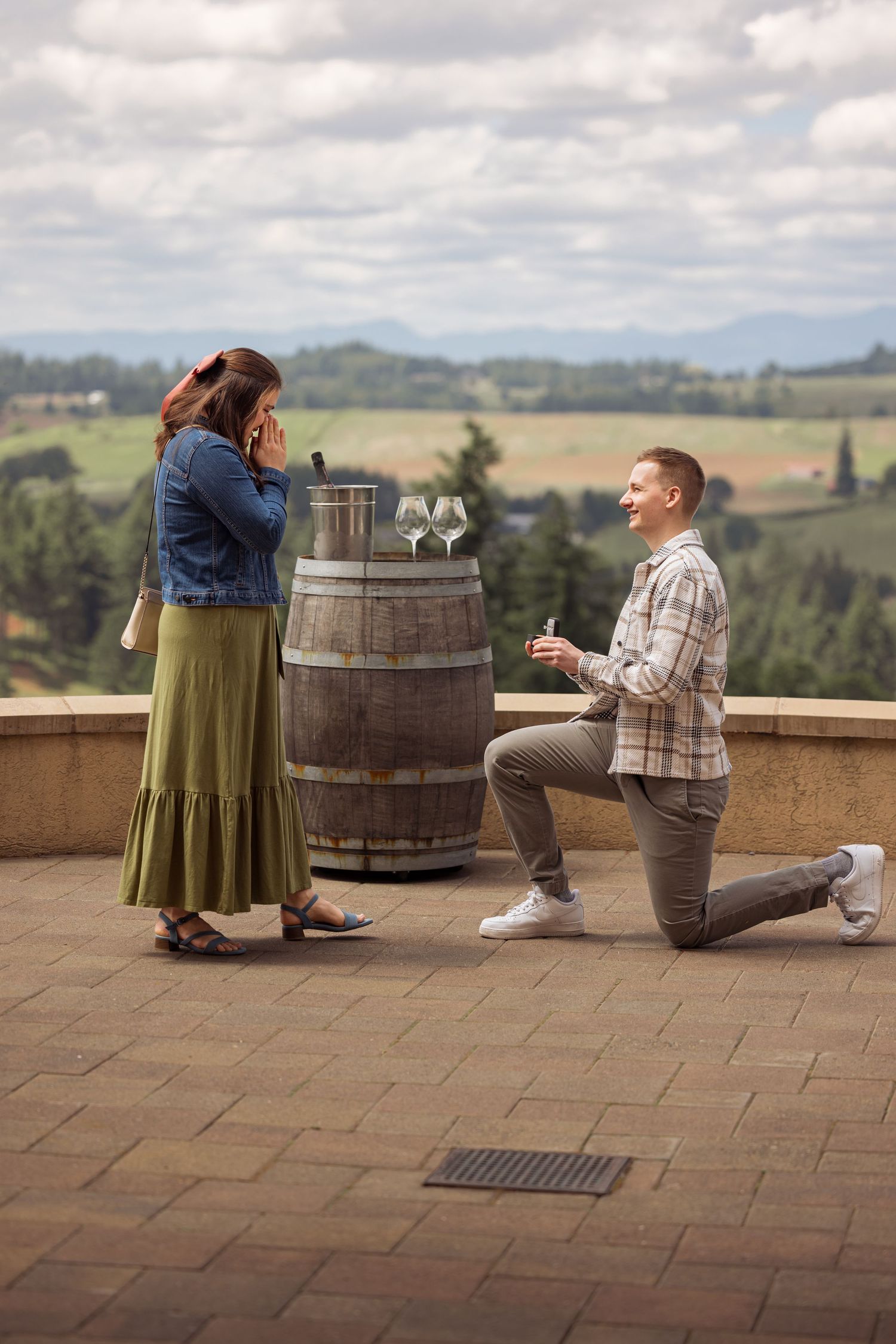 Man proposes to woman on one knee at scenic winery terrace with wine glasses on barrel and rolling hills backdrop.