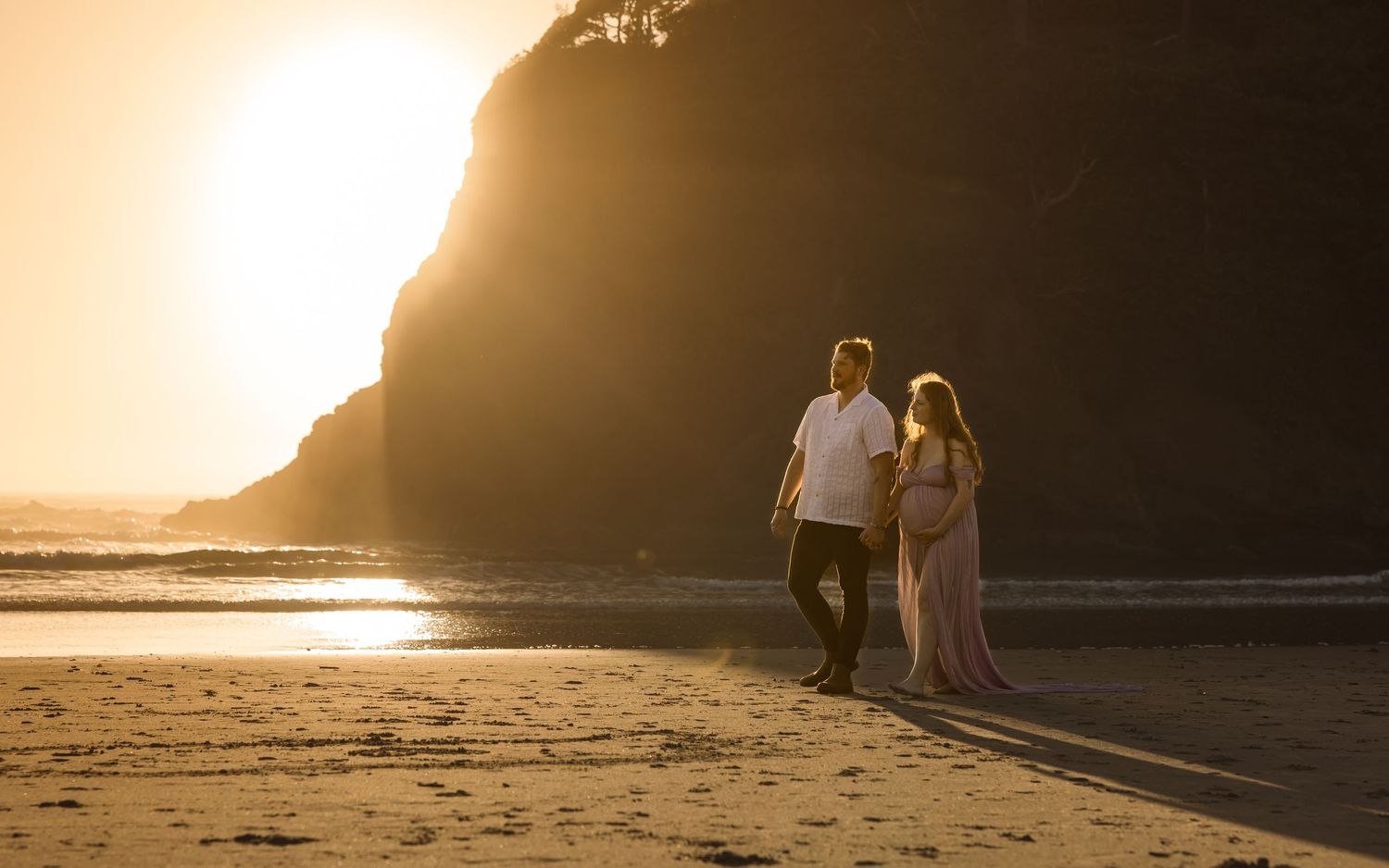 A couple strolls along a beach at sunset with a dramatic cliff silhouetted against the golden sky.