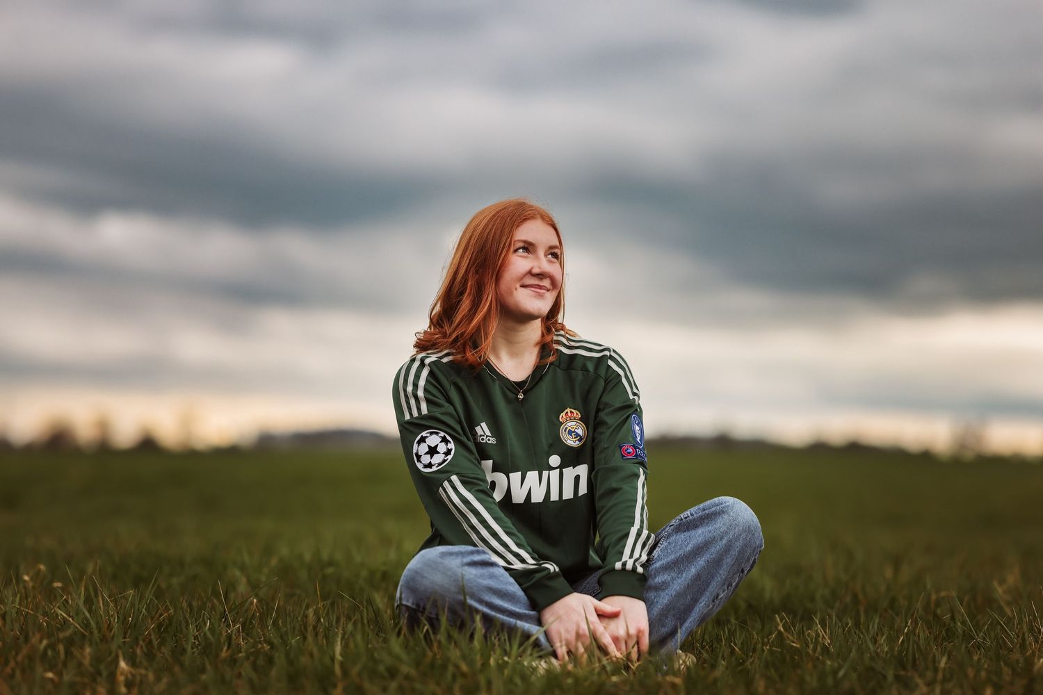 Person in jersey sitting in a grassy field under dramatic cloudy sky during a photo session taken by Salem, Oregon senior photographer.