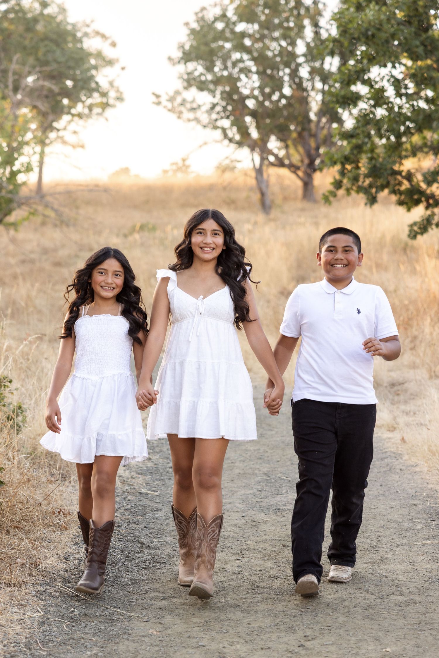 Three siblings in white outfits walk hand-in-hand on a dirt path surrounded by golden grass and trees.
