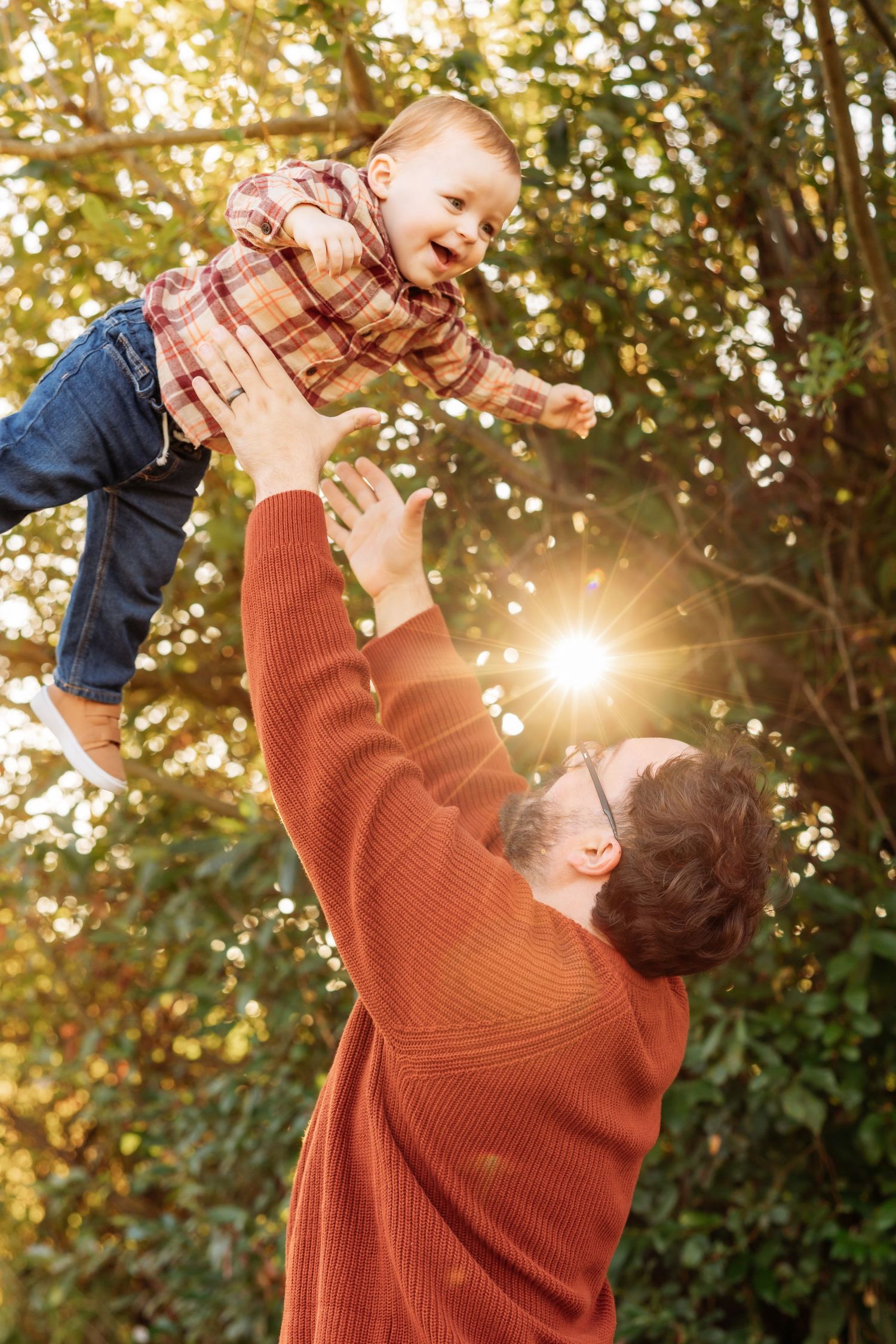 Father joyfully lifting laughing baby in autumn park with golden sunlight streaming through trees.