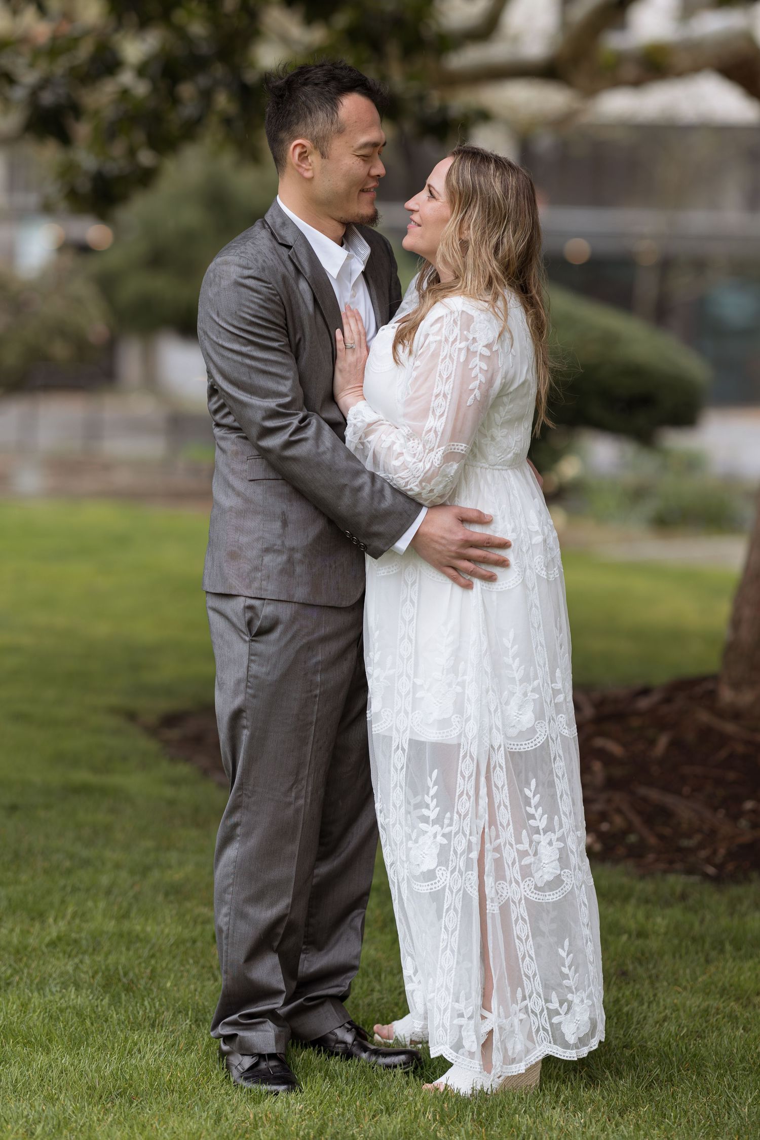 Couple in wedding attire embracing outdoors, man in gray suit, woman in white lace dress, smiling at each other.