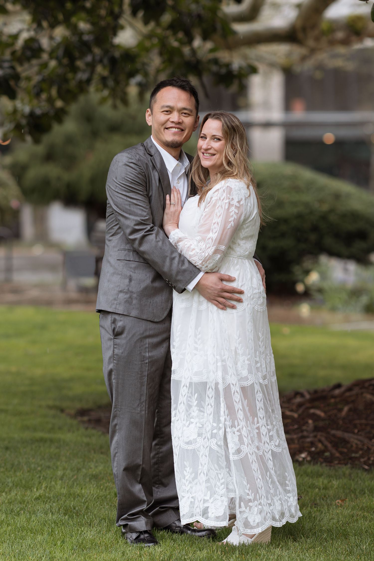 Happy couple embracing outdoors, woman in white lace wedding dress and man in gray suit, smiling on green lawn.