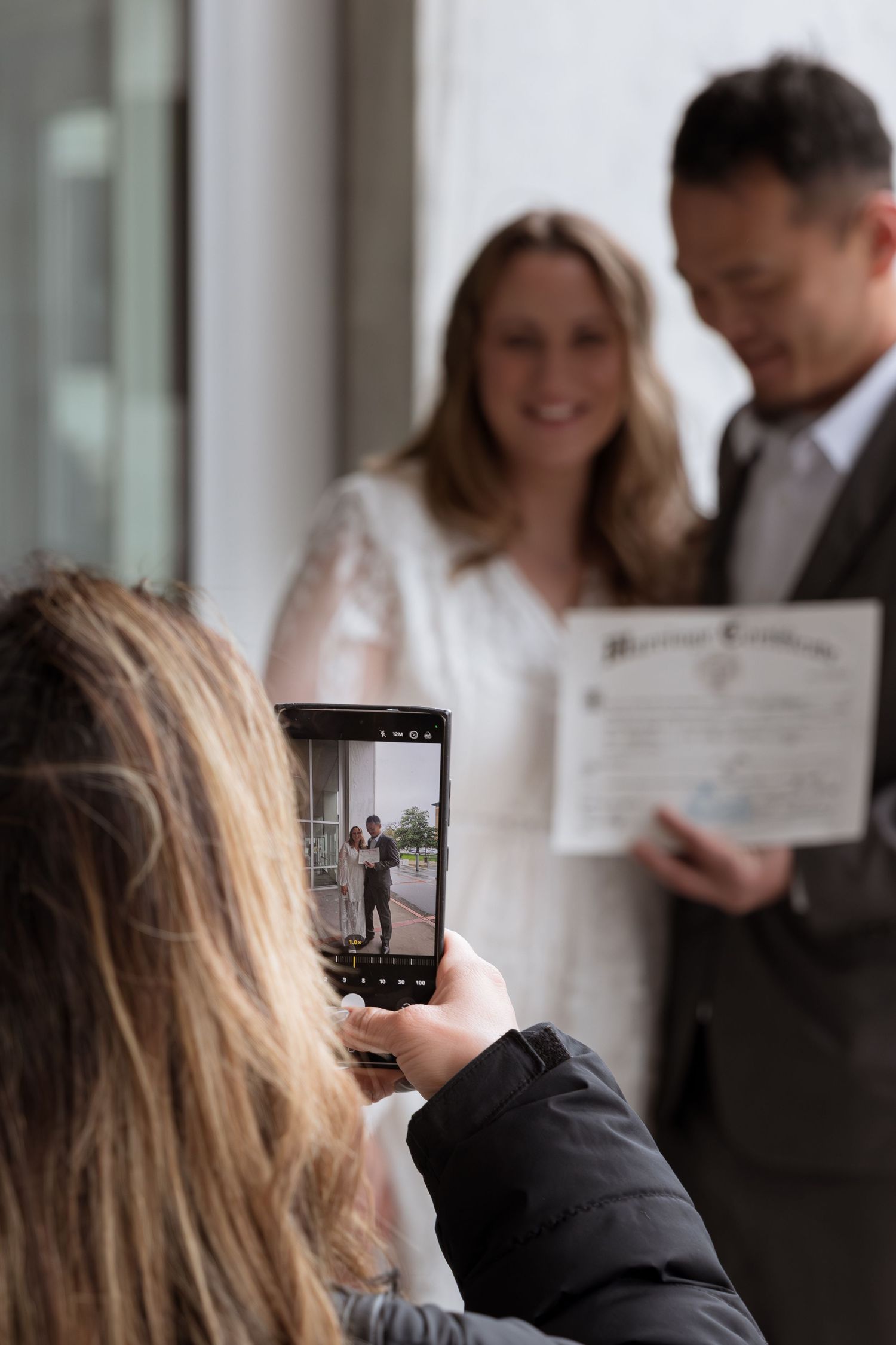 Person photographing a couple holding a marriage certificate on a smartphone at a wedding ceremony.
