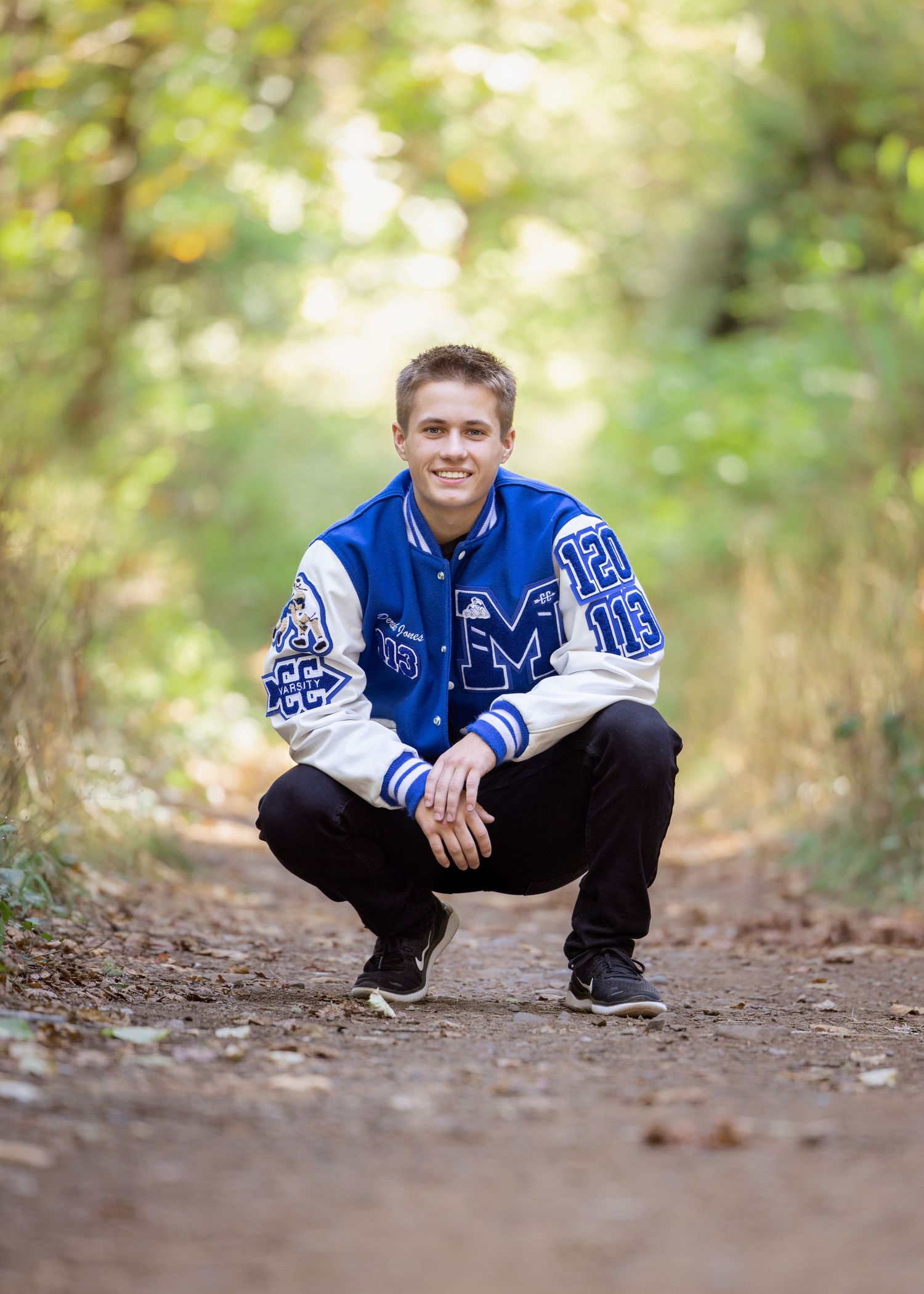 Student in varsity letterman jacket crouches on a forest path during a photo session taken by Salem, Oregon senior photographer.