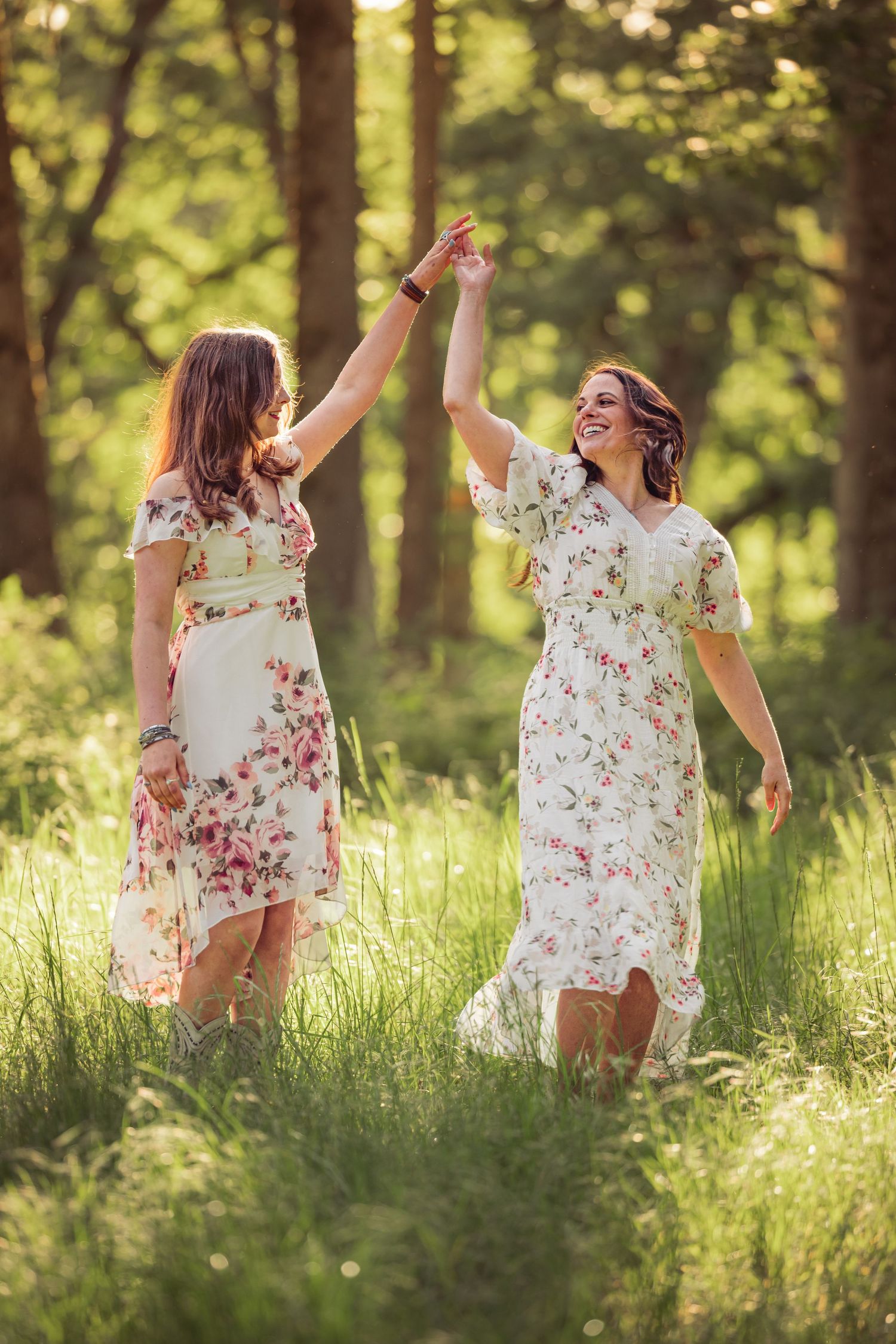 Two women in floral summer dresses dance together joyfully in a sunlit forest clearing with tall grass.