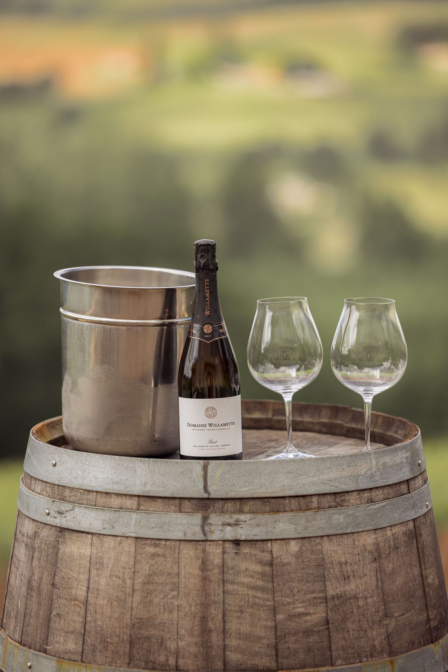 Champagne bottle in ice bucket with two wine glasses arranged on rustic wooden wine barrel outdoors.