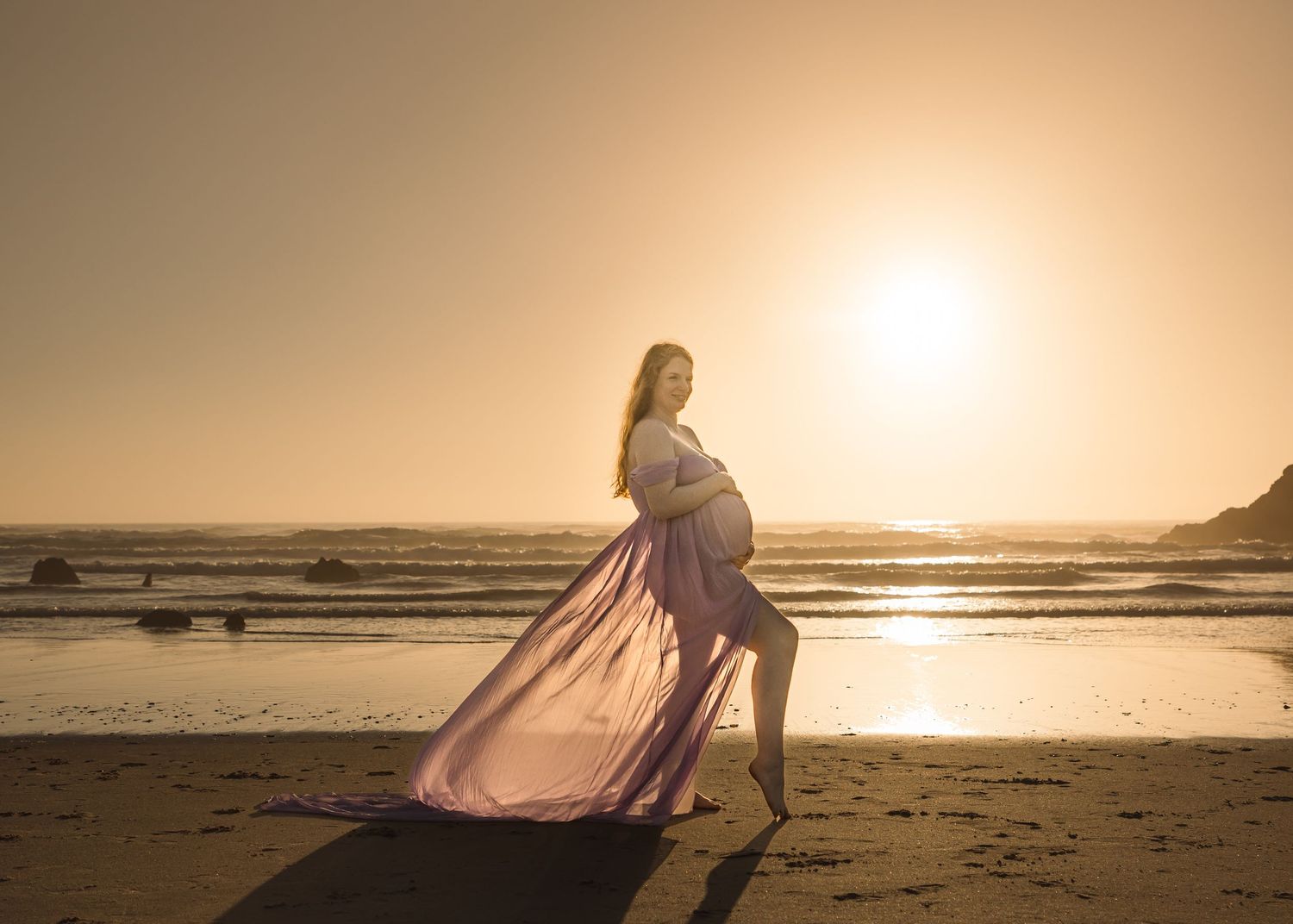 Silhouette figure in flowing dress stands on beach at golden sunset with ocean waves in background.