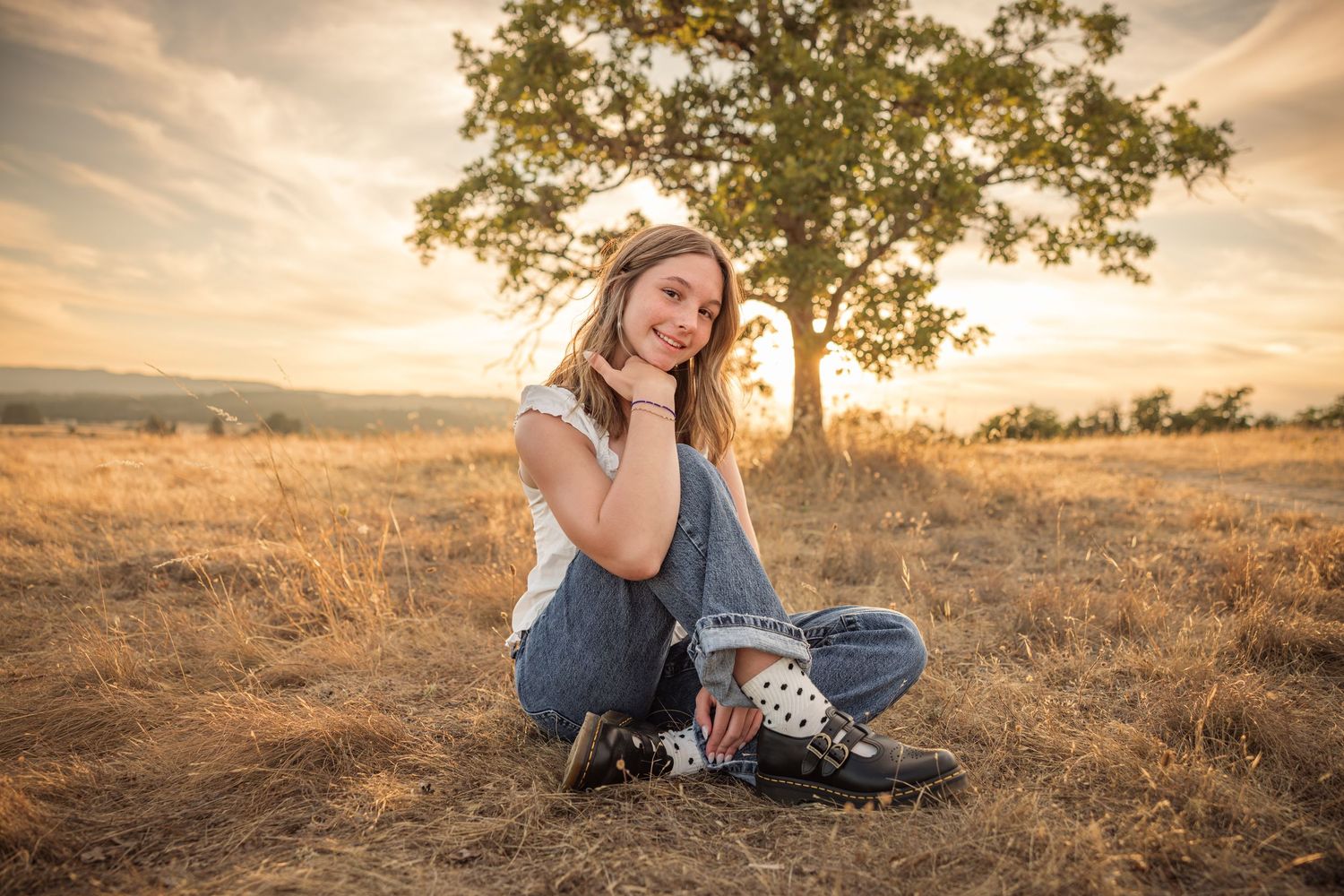 Young woman sitting cross-legged in golden field at sunset, smiling, with a tree and warm sky behind her.