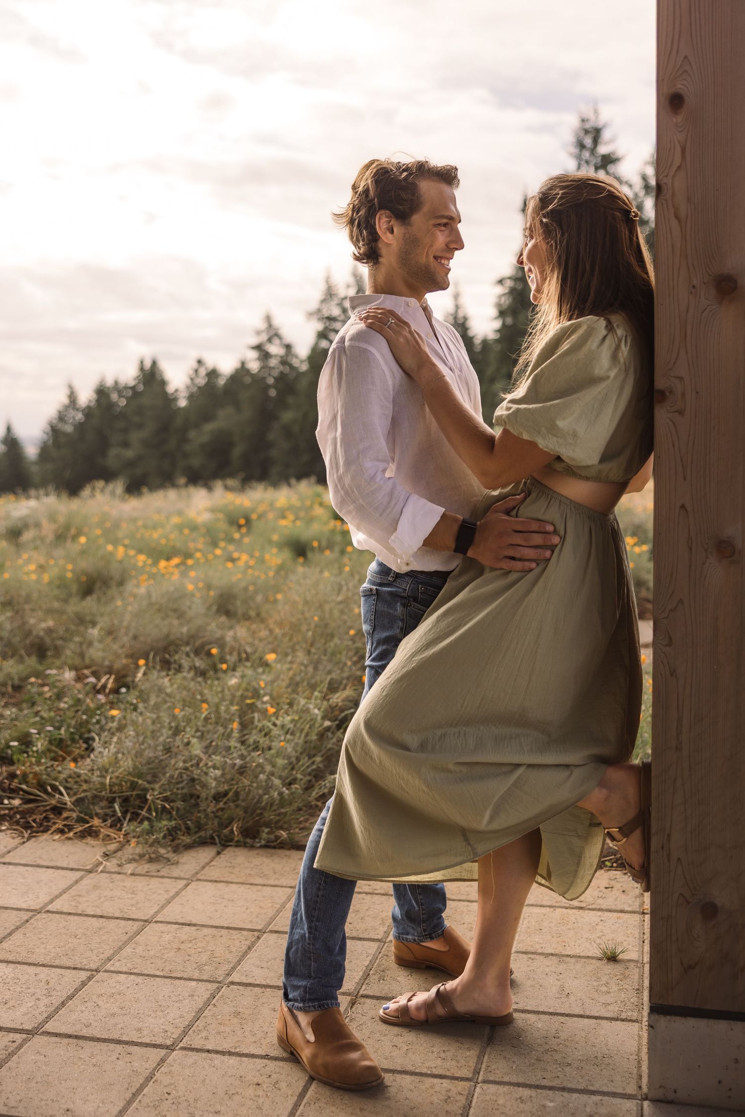Two people share an intimate moment sitting together in a rustic outdoor setting with wildflowers in the background.