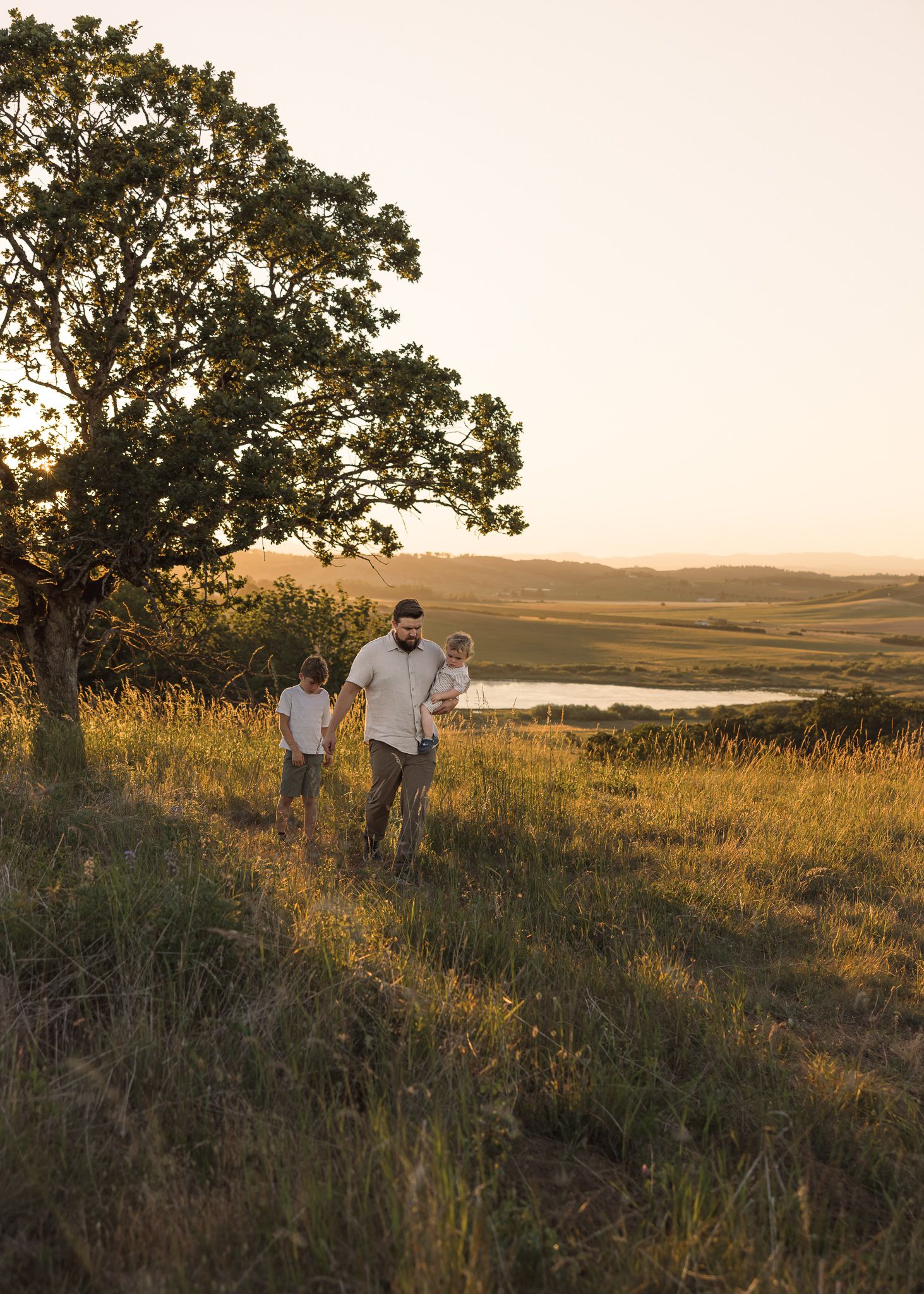 Family stands on a grassy hillside overlooking rolling fields at sunset with a large tree nearby taken by Salem family photographer.