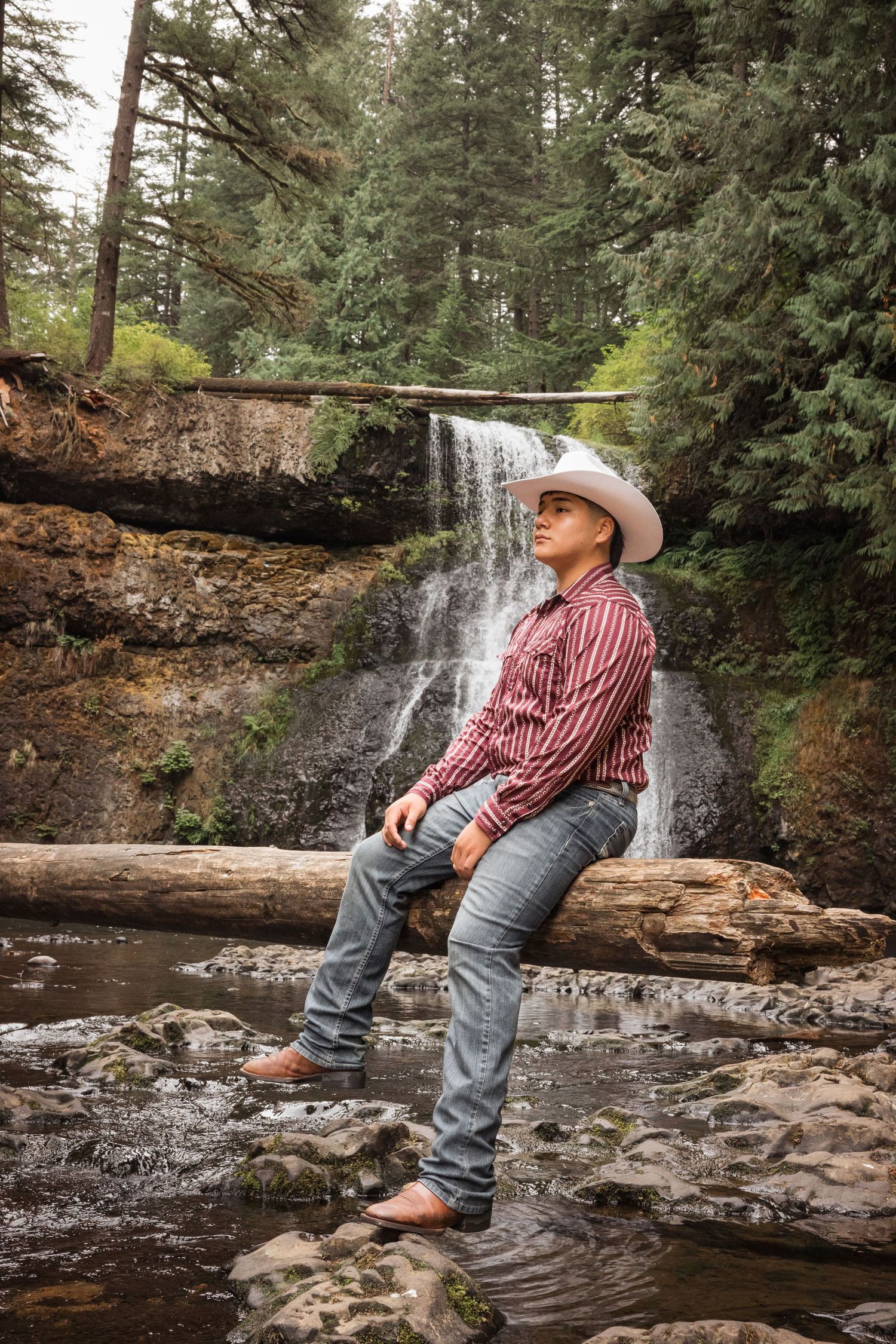 A rustic outdoor portrait session by a cascading waterfall featuring someone in a striped shirt and cowboy attire.