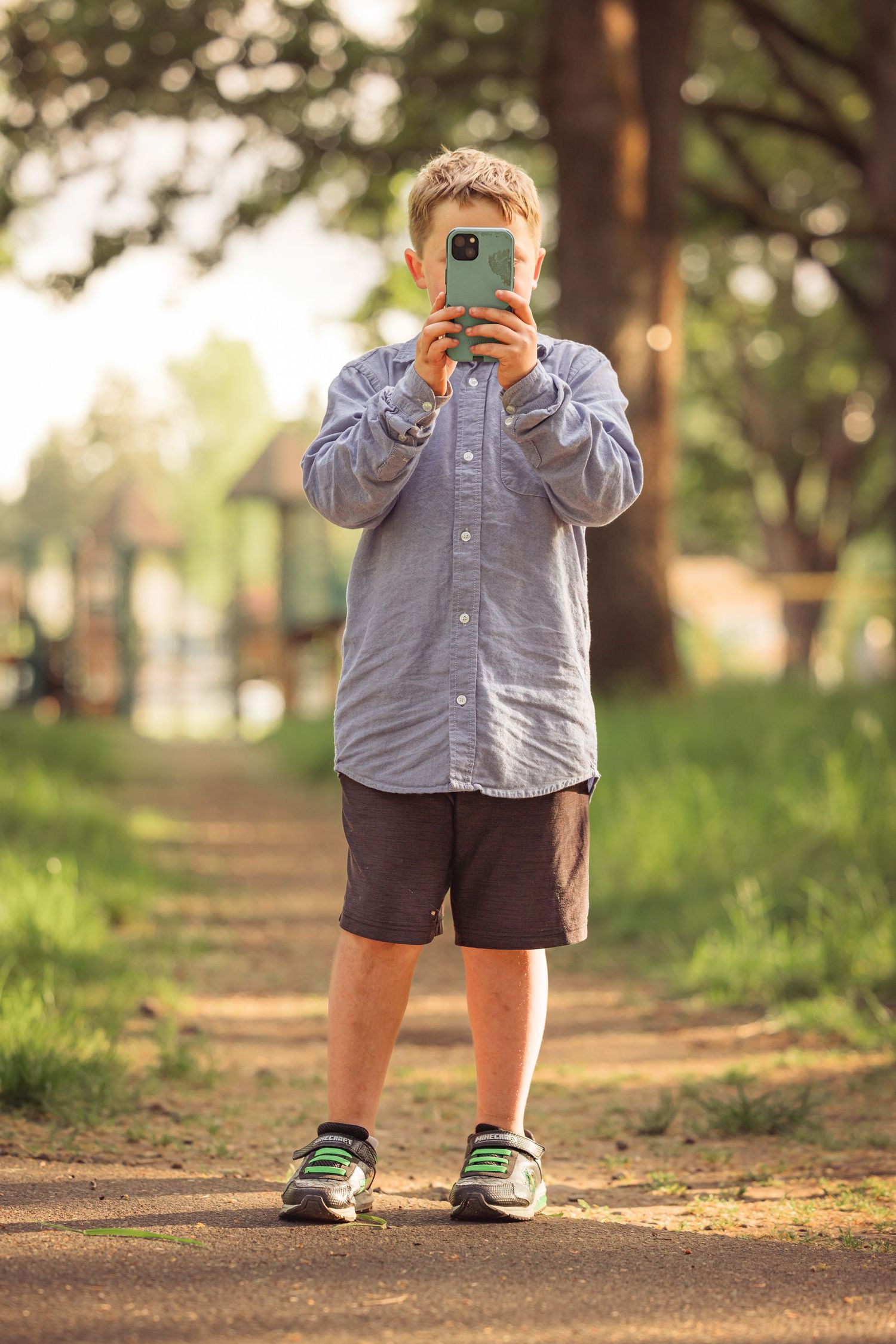 A person wearing a gray button-down shirt and shorts photographs with a mobile phone on a tree-lined dirt path.