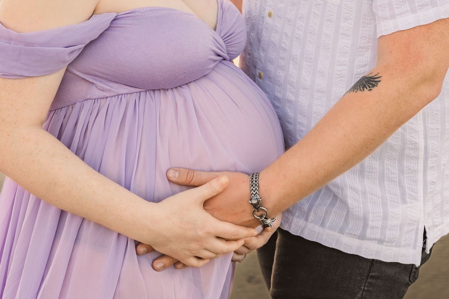 Hands gently rest on a pregnant belly draped in a flowing lavender maternity dress beside someone in a white shirt.