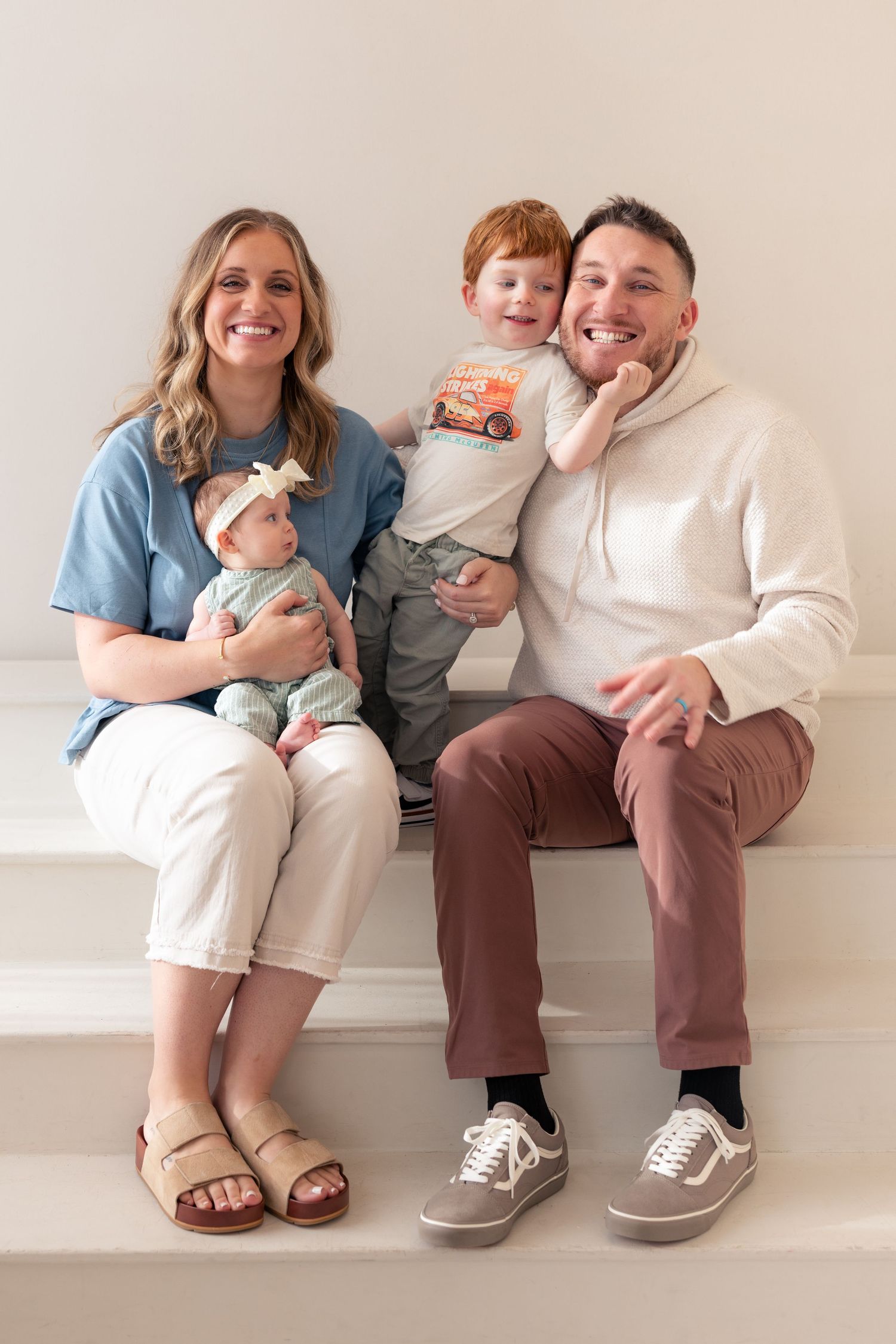 Happy family of four sitting on white stairs, parents holding a toddler and newborn baby.