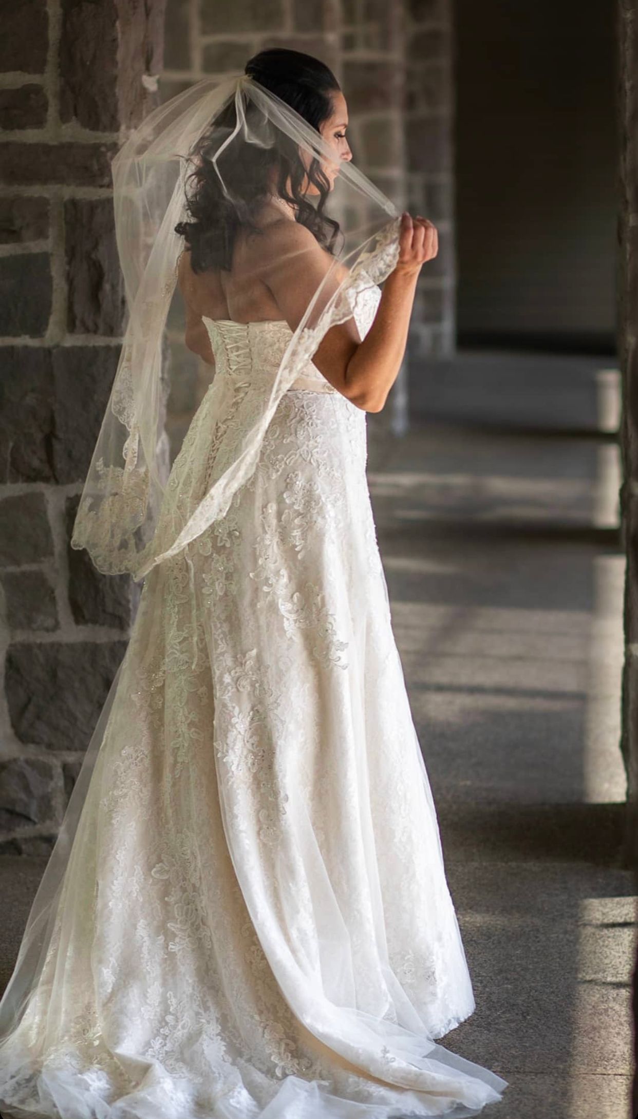 Bride in strapless lace wedding gown with flowing veil, standing in profile against stone wall.