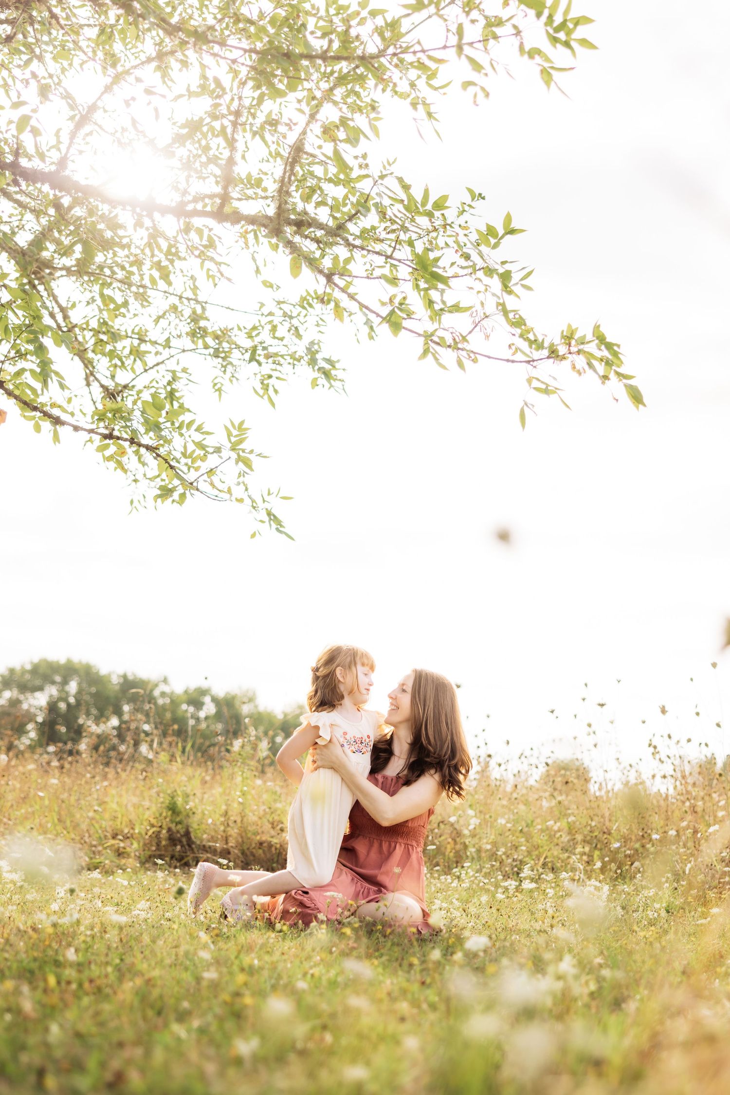 A mother and young daughter share a tender moment sitting in a sunlit meadow beneath a blossoming tree.