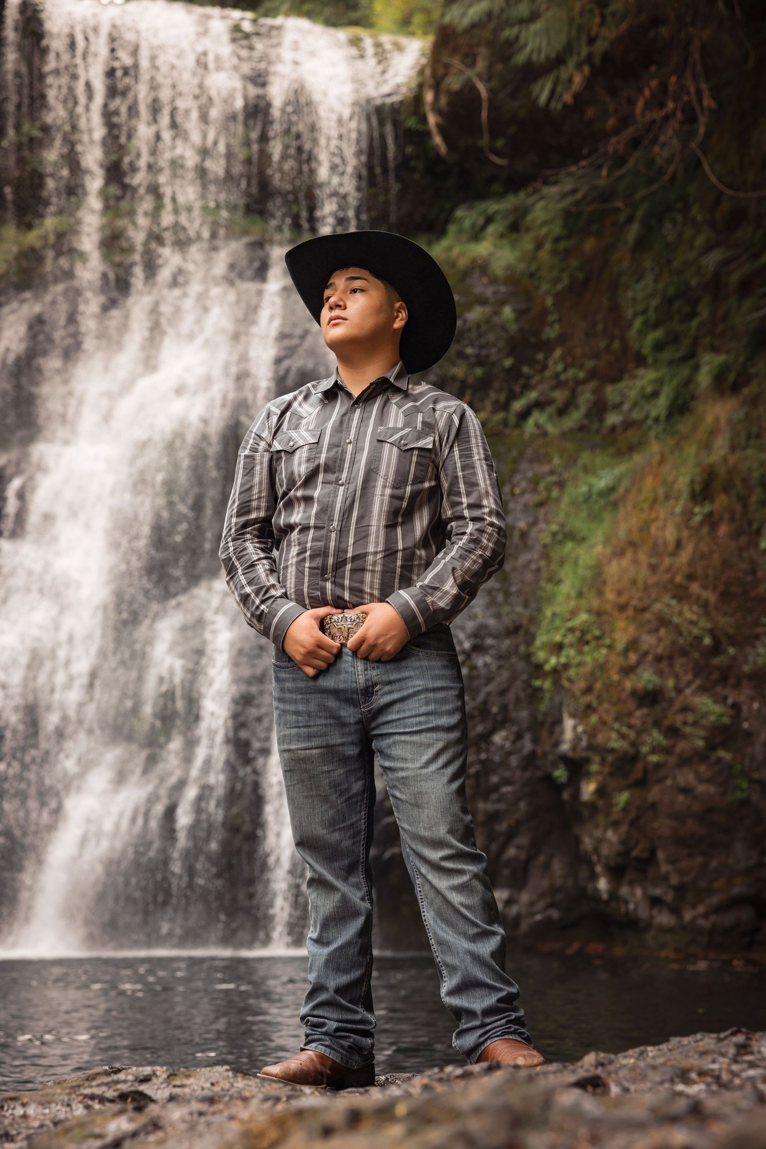A scenic photoshoot at a majestic waterfall with a person wearing western attire and a black cowboy hat.