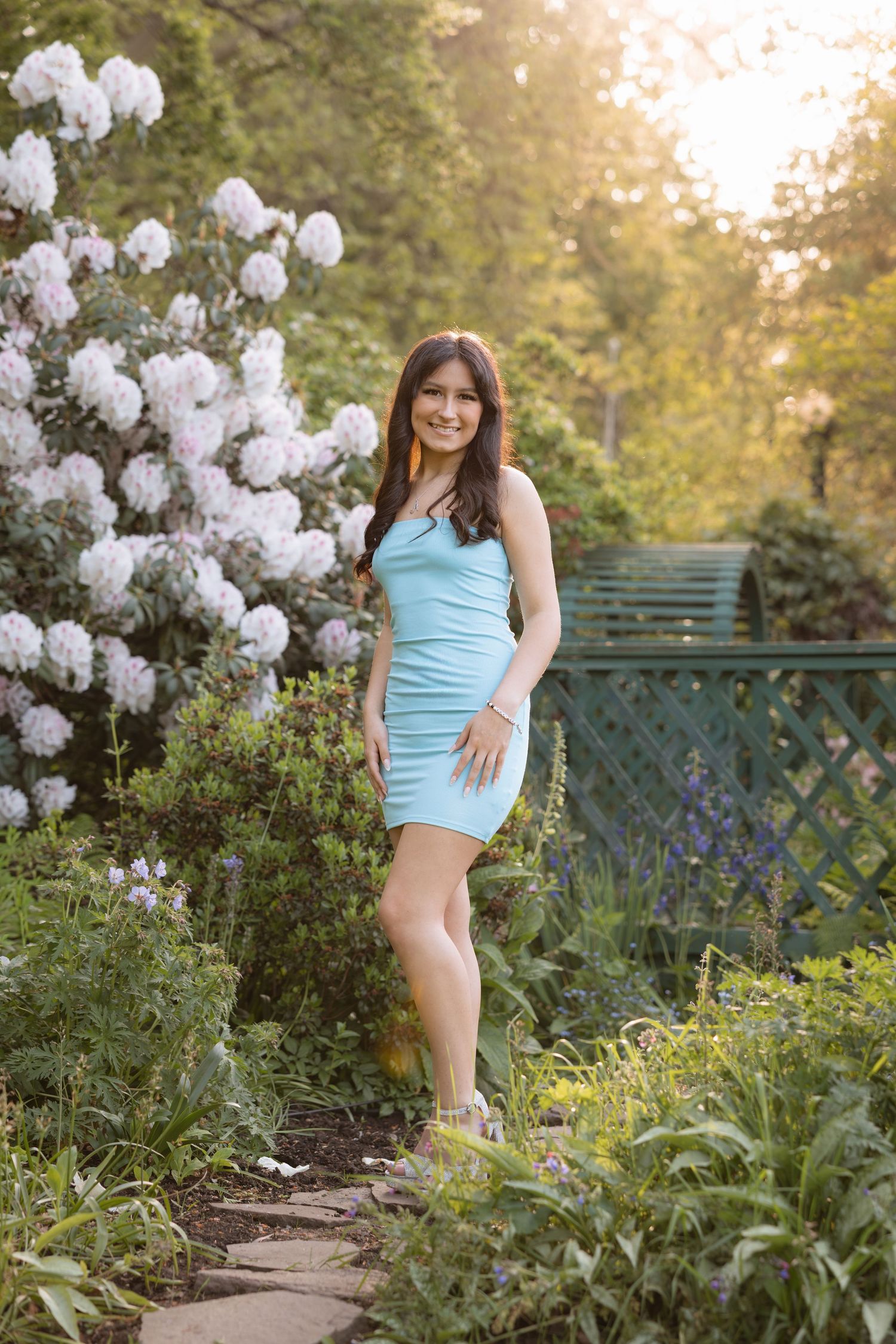 Young woman in light blue mini dress posing in a lush garden with white flowering bushes at golden hour.